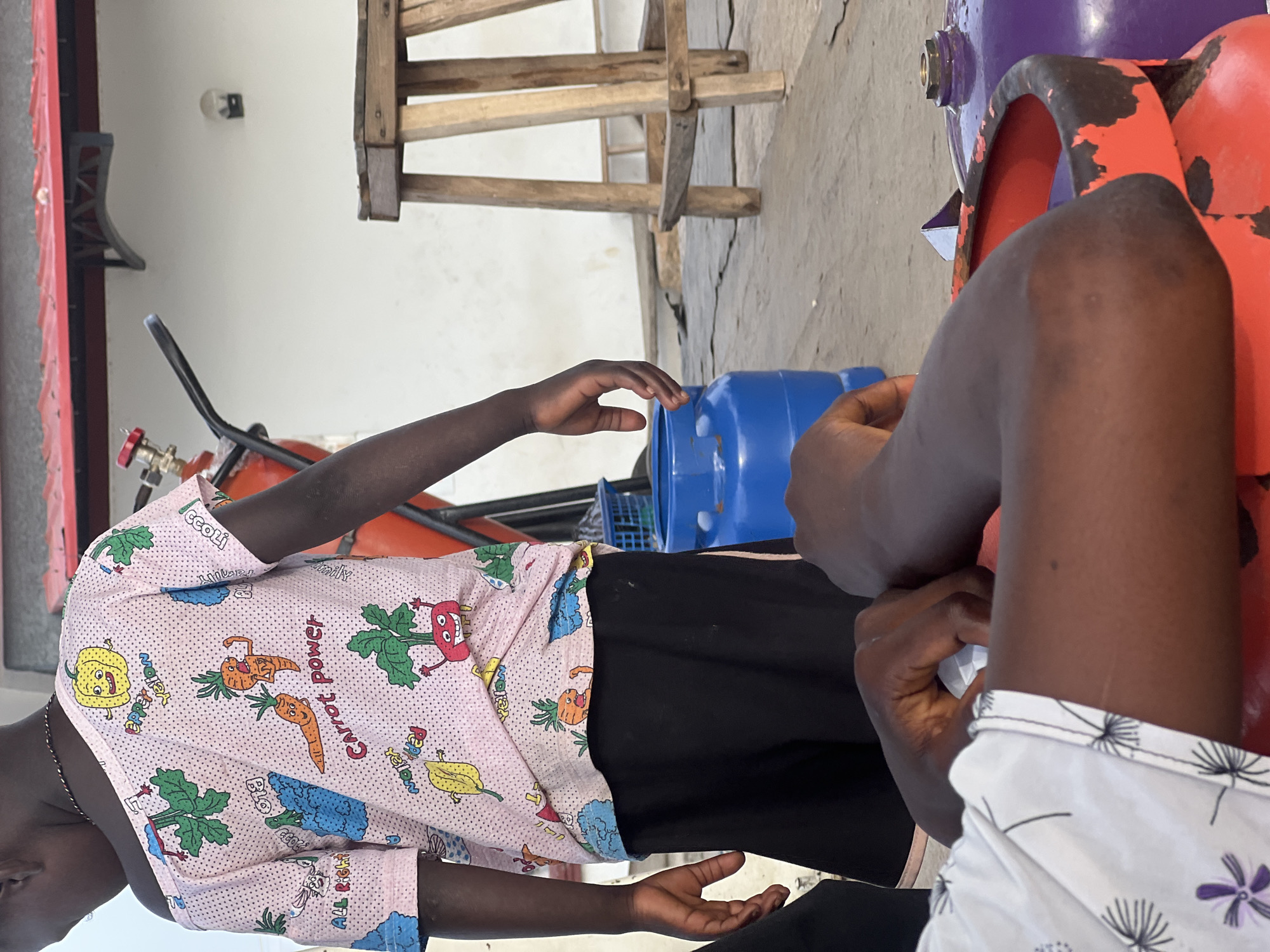 The image depicts a scene likely on a rooftop or open-air space in Jalingo, Nigeria. In the foreground, an individual's arms are visible, with one hand resting on a large red gas cylinder that has a purple gas cylinder next to it. Both cylinders appear to be in use or recently used. Further back, a child, wearing a pink shirt with cartoon carrot and vegetable prints, stands with their back to the camera. The child is wearing black shorts. Behind the child, a large red propane tank with a visible valve is situated. A blue plastic basket and a blue gas cylinder are also present. In the background, a white wall with a red-tiled roof and a wooden stool can be seen. The overall impression is one of a domestic or semi-industrial outdoor setting.