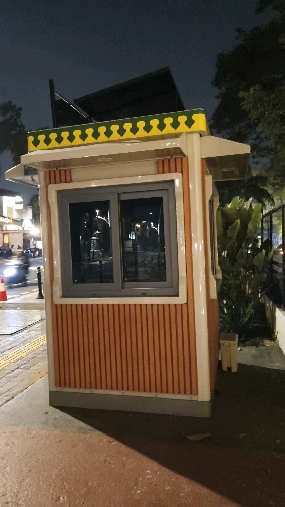 A night-time outdoor scene in South Jakarta, Indonesia, features a small, brightly colored guard booth. The booth has a beige and orange striped exterior with a decorative yellow and green trim along the roofline. A large, reflective window is visible on the front of the booth. To the left, a traffic cone sits on a sidewalk next to a street with blurred car lights, indicating movement. In the background, trees and foliage are visible, along with some buildings and streetlights. The ground in front of the booth appears to be paved with reddish-brown material.