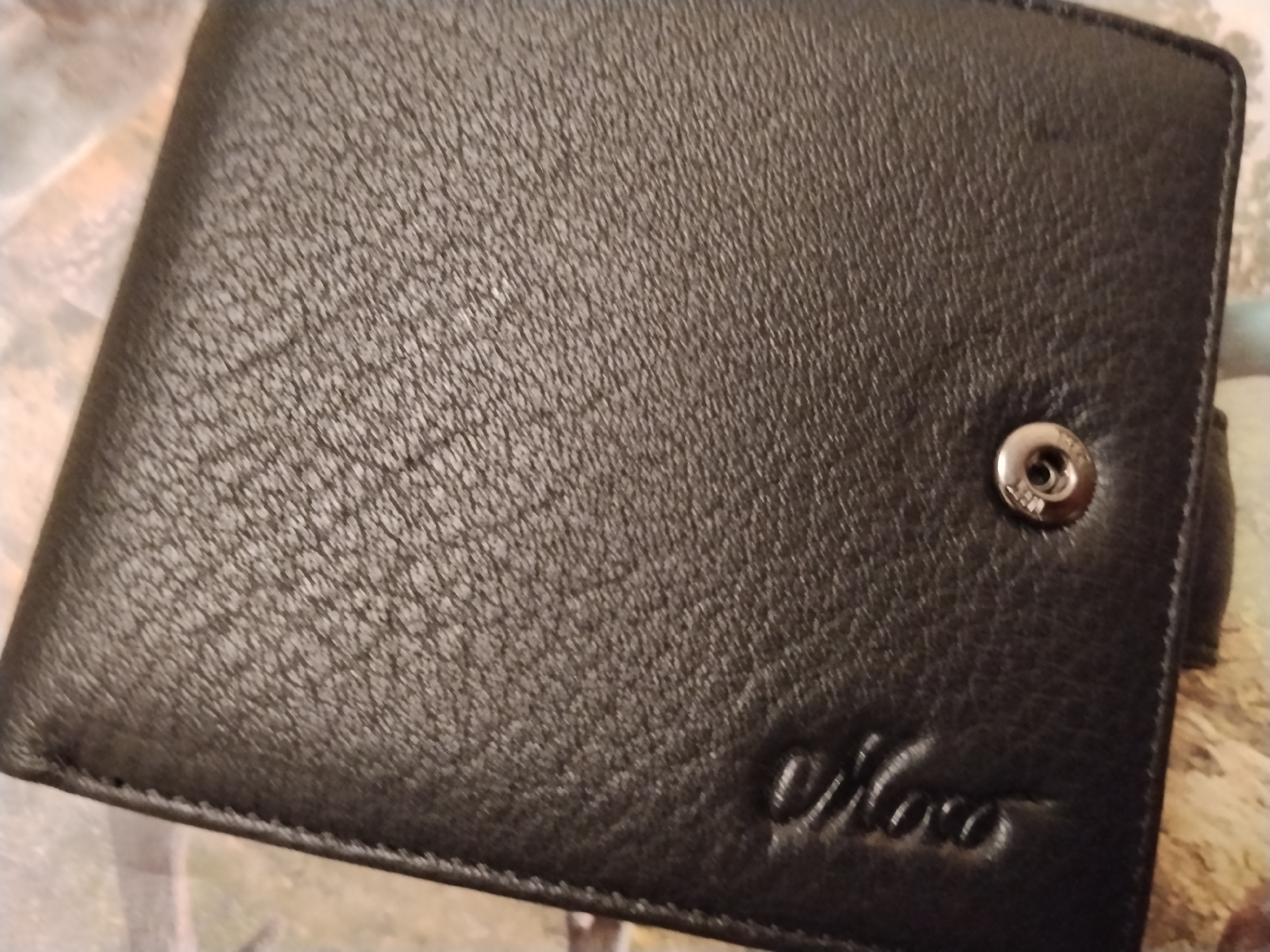 The image shows a close-up, overhead view of a dark brown, textured leather bi-fold wallet. The wallet is lying flat. A silver snap closure with an embossed logo is visible on the right side. Embossed in the bottom right corner of the wallet is a cursive logo that appears to read "Noco". The background is a light-colored, patterned surface. There are no people, actions, or interactions depicted. The location is unknown, and no specific details about Iziaslav, Ukraine are visible.