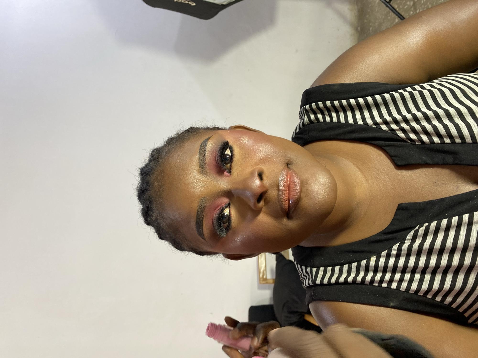 The image shows a woman with dark skin and dark hair, wearing a black and white striped sleeveless top. She has makeup on, including red eyeshadow and lip gloss. A hand is visible in the foreground, holding a pink object. The background is a plain white wall. The location is Gura Top, Nigeria.
