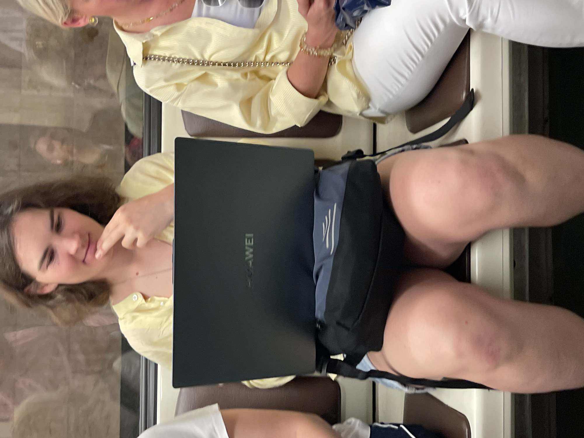 In Moscow, Russia, on what appears to be a public transport vehicle such as a metro train, a young woman with light brown hair, wearing a light yellow shirt, sits with a laptop open on her lap. A dark blue bag is placed beneath the laptop. Her right hand is raised to her chin, with her index finger touching her lips, as she looks down at the screen. To her right, another woman, with blonde hair and wearing a light yellow shirt and white pants, is seated, looking forward. The background shows reflections in a window, including blurred figures of other passengers. The seats are upholstered in brown and white.