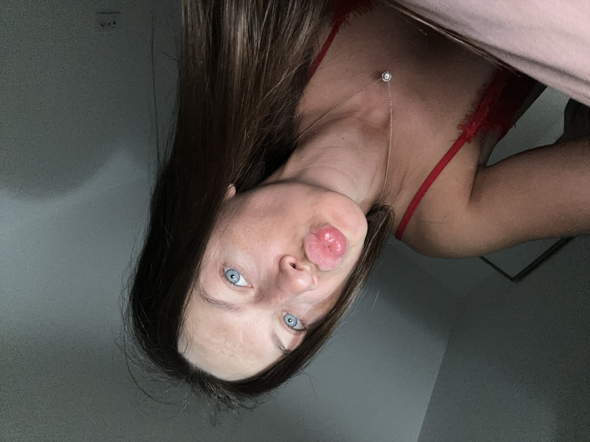A young woman with long brown hair and bright blue eyes is captured in a close-up, selfie-style shot. She is wearing a red bralette and a delicate silver necklace with a single diamond pendant. Her lips are puckered as if blowing a kiss. The background appears to be an indoor setting, with a slanted ceiling and muted lighting, suggesting a bedroom or similar private space. The woman's skin shows some natural texture and subtle markings, indicating a candid and unposed moment. The overall impression is one of casual intimacy. There is no information about the location in Donji Pobori, Montenegro, within the image itself.