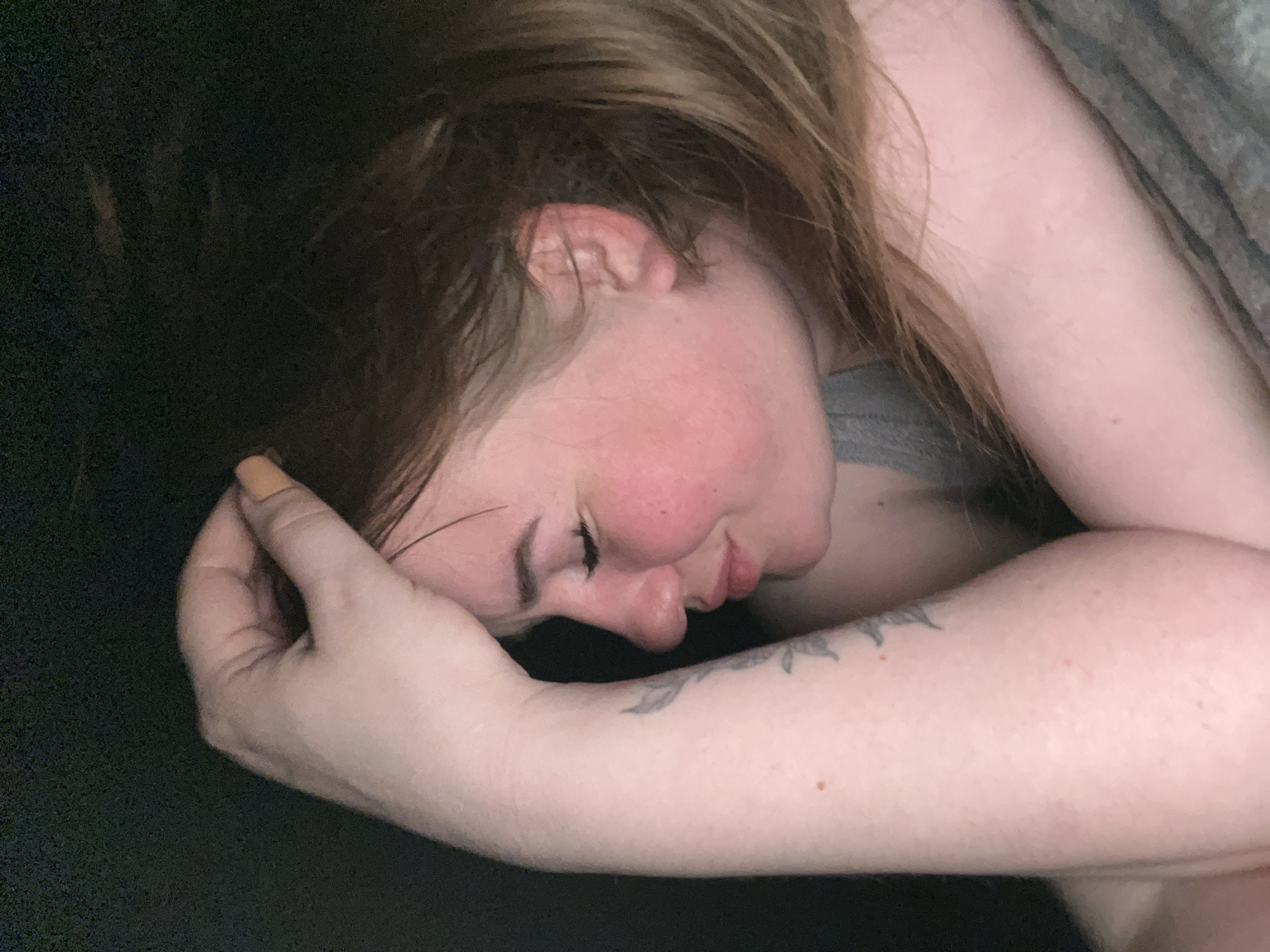 A close-up shows a woman lying down. Her head is turned to the side, and her eyes are closed. Her right arm is bent and positioned near her head, with her hand near her face. She has light skin, reddish-brown hair, and a tattoo on her left arm. The environment is dimly lit, with some dark fabric visible in the background. There is no evidence to suggest the location is Chaikovsky, Russia.