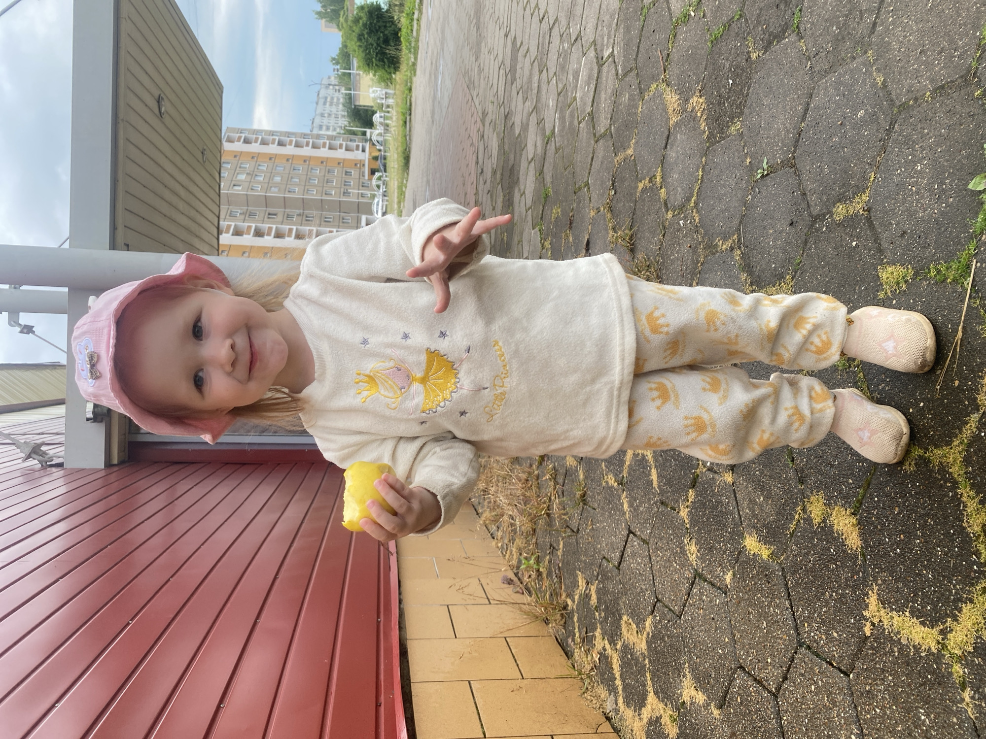 A young girl wearing a pink hat and a light-colored pajama set with an elephant pattern is standing on a paved walkway. She is holding a half-eaten yellow apple in her left hand and has her right hand raised with her fingers spread. The girl is looking at the camera and smiling. The background shows a red building on the left and apartment buildings in the distance, suggesting an urban or suburban residential setting. The walkway is made of interlocking hexagonal and rectangular concrete pavers, with patches of grass growing between them. The sky is visible in the upper part of the image, appearing overcast or cloudy. The overall impression is of a casual outdoor moment, possibly during daylight hours. Text is visible on the girl's pajama top, appearing to read "Little Princess."
