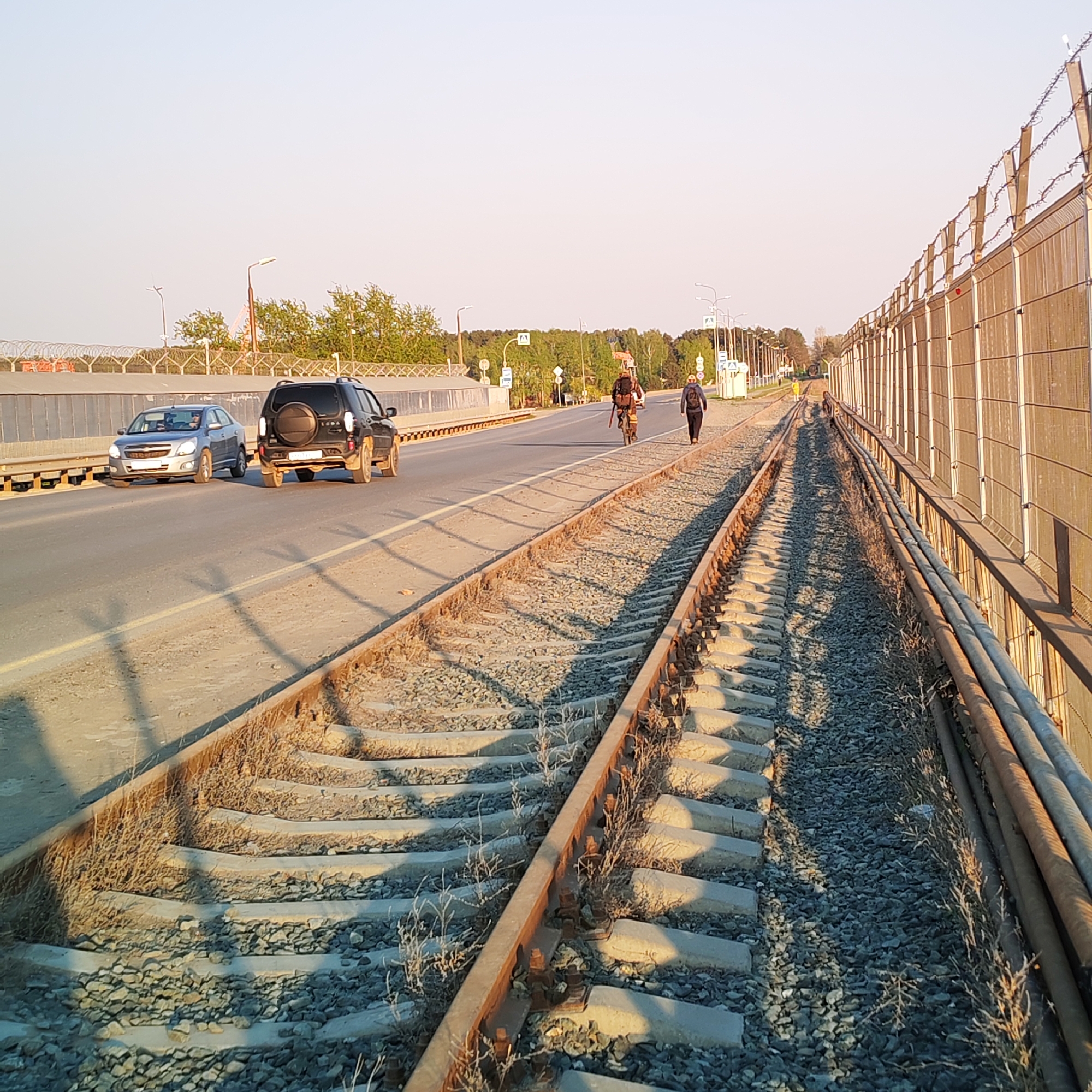 The image depicts a scene in Chaikovsky, Russia, featuring a section of railway tracks running parallel to a road on a bridge. Two cars, a silver sedan and a black SUV, are traveling on the road. Further down the road, a person is cycling, followed by another person walking. A security fence with barbed wire is visible along the right side of the railway tracks. The railway tracks themselves show signs of disuse with vegetation growing around the ties and gravel bed. Overhead, there are street lights and a pedestrian crossing sign. The scene is bathed in the soft light of either early morning or late afternoon.