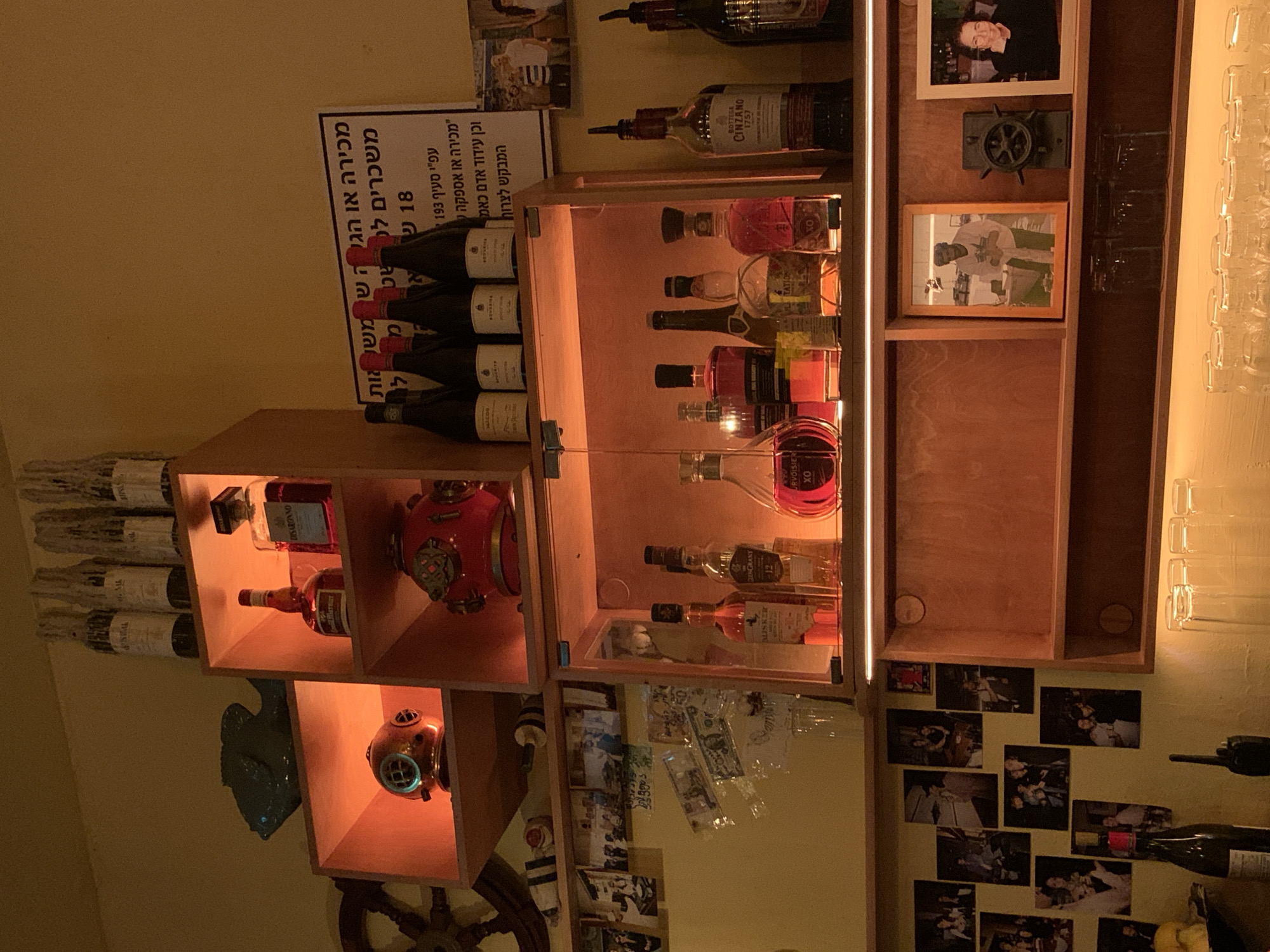 The image shows the interior of a bar or restaurant. It appears to be a bar in Tel-Aviv, Israel based on the Hebrew writing on a sign next to the wines.
The scene consists of a wooden display case with shelves. The shelves contain bottles of wine, liquor, and decorative nautical-themed objects, such as red diving helmets. There are also framed photographs and an antique ship's wheel. The wall behind the display is covered in a collage of black and white photographs and currency notes.