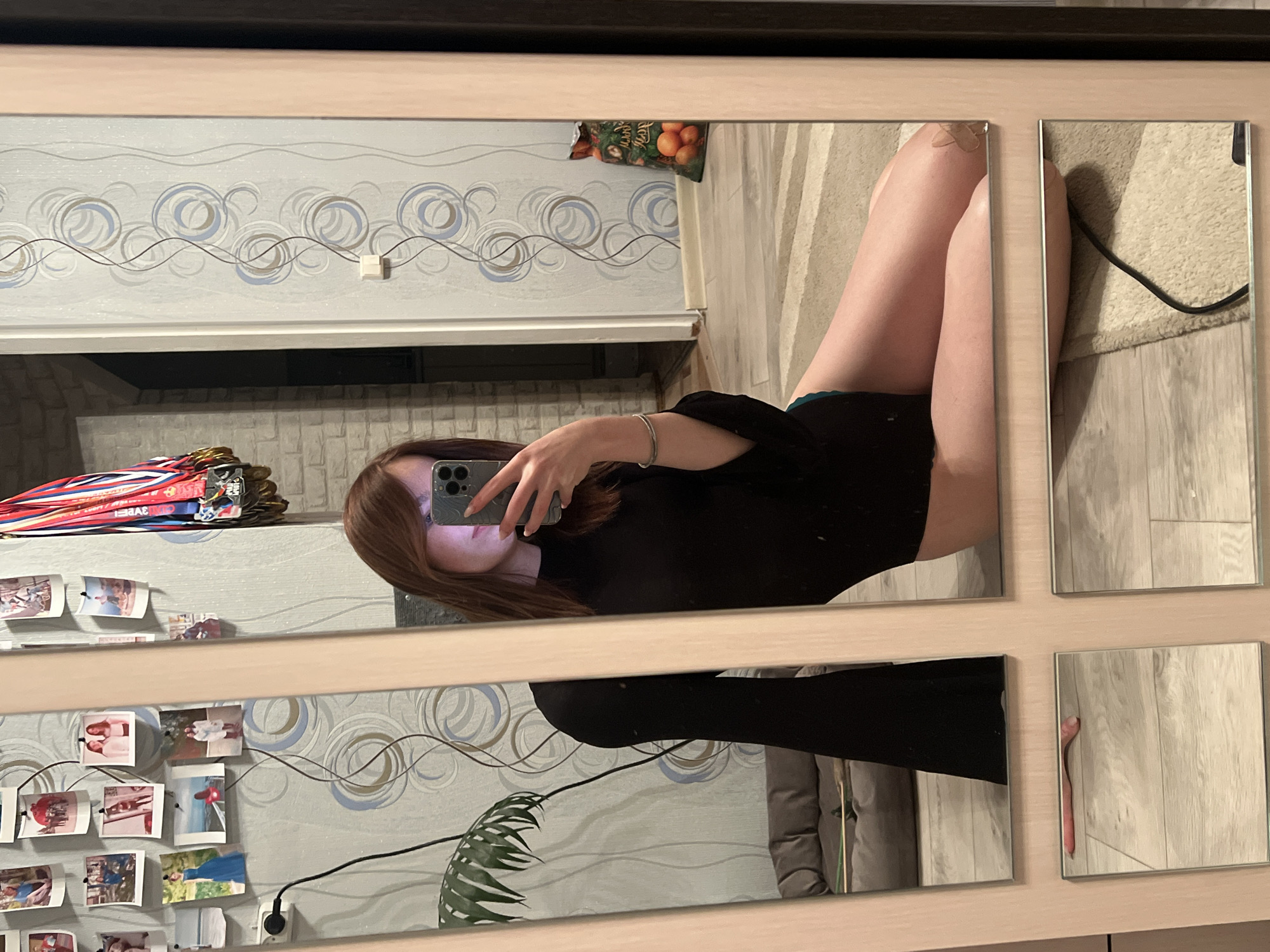 A young woman is depicted in a mirror selfie, kneeling or sitting on a light-colored rug over a wooden floor within an indoor residential setting in Chaikovsky, Russia. She wears a black long-sleeved, high-necked top, possibly a bodysuit, and green underwear. Her reddish-brown hair frames her partially obscured face as she holds a dark-colored smartphone, likely an iPhone, with her right hand to capture her reflection. A silver-toned bracelet is visible on her left wrist, and a small, beige bandage is adhered to her right knee. The wall behind her features light grey wallpaper patterned with blue and grey concentric circles and vertical wavy lines. This wall is decorated with multiple polaroid-style photographs and a display of sports medals featuring red, white, and blue ribbons. To the far left of the reflection, a grey cushioned sofa or pet bed is partially visible, along with green foliage from a houseplant. To the right, a patterned sack filled with oranges rests on the floor. A white electrical outlet is present on the patterned wall. A black electrical cable lies across the rug, and a pink object, possibly a hairbrush, is visible on the wooden floor reflected in a lower mirror panel.