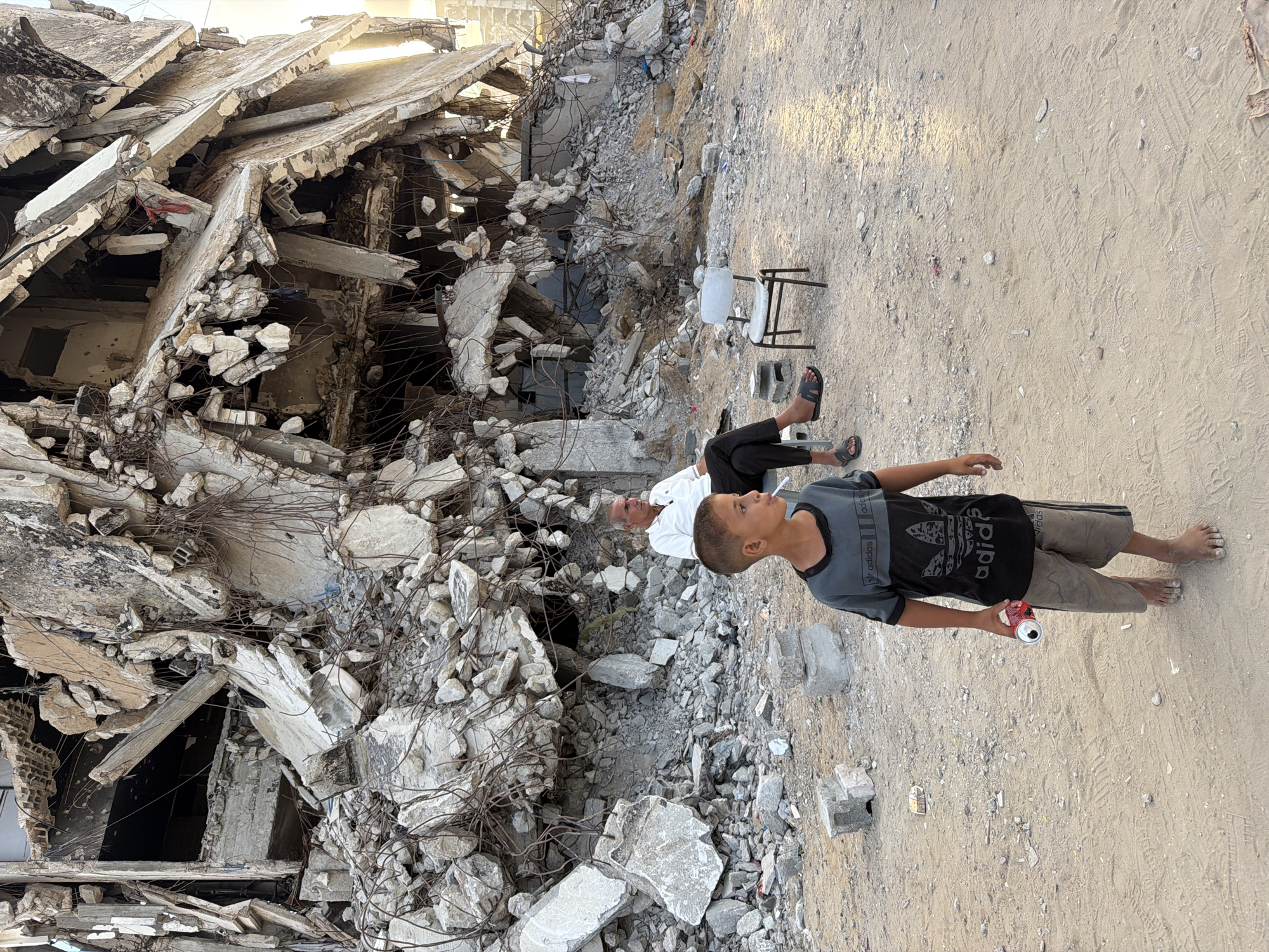 An adult male wearing a white polo shirt and dark pants sits in a chair near a demolished building in Gaza City, Palestinian Territories. A young boy, barefoot and wearing a grey and black t-shirt and khaki shorts, stands in the foreground, looking to his left and holding a red and white object. The background shows the heavily damaged remains of a multi-story building, with exposed rebar and rubble scattered on the ground. Another chair is placed near the adult male, and several concrete blocks are visible on the sandy ground. The lighting suggests it is either early morning or late afternoon.