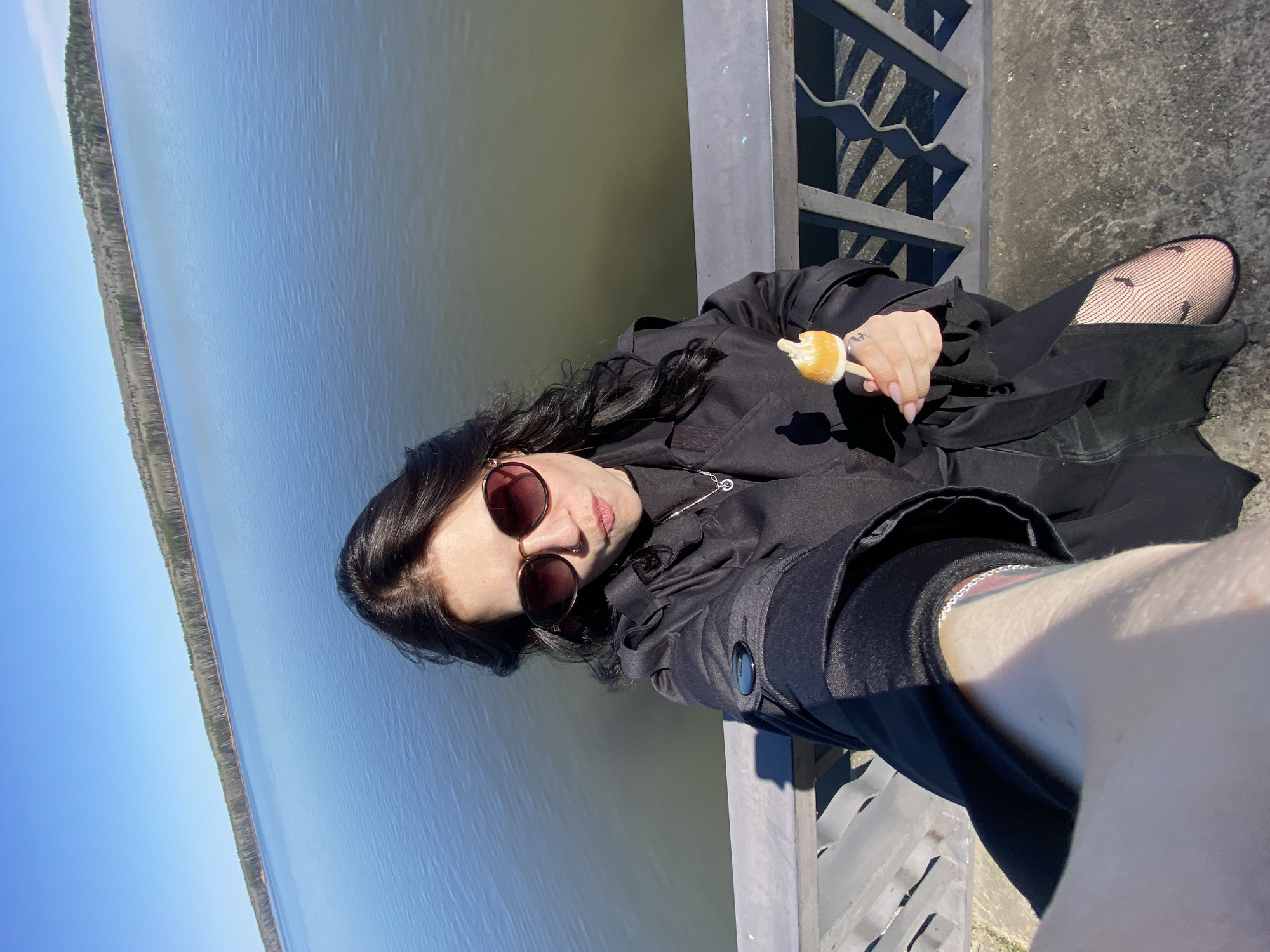 A woman is holding a popsicle while sitting on the edge of a bridge in the city of Chaikovsky, Russia. She is wearing sunglasses, a dark jacket, and patterned tights. A body of water with trees lining the far shore is visible in the background, with a blue sky overhead. The woman appears to be taking a selfie.