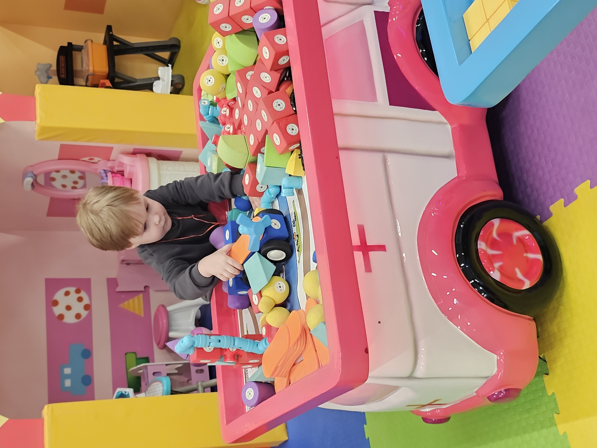 A young, light-haired child is interacting with a large, colorful toy vehicle, likely an ambulance or similar service vehicle, filled with various play blocks and shapes. The vehicle has a pink frame, a white body with a red cross symbol, and blue and pink accents. The child, wearing a grey sweater, is reaching into the vehicle, presumably playing with the blocks.
The scene is set in an indoor play area, recognizable by the colorful foam mats and playful decorations on the walls. The walls are adorned with pink and white stripes, polka dots, and cartoon car decals. In the background, there are additional play structures and equipment.
It's possible this scene is located in a play area or children's entertainment center in the city of Khimki, Russia.