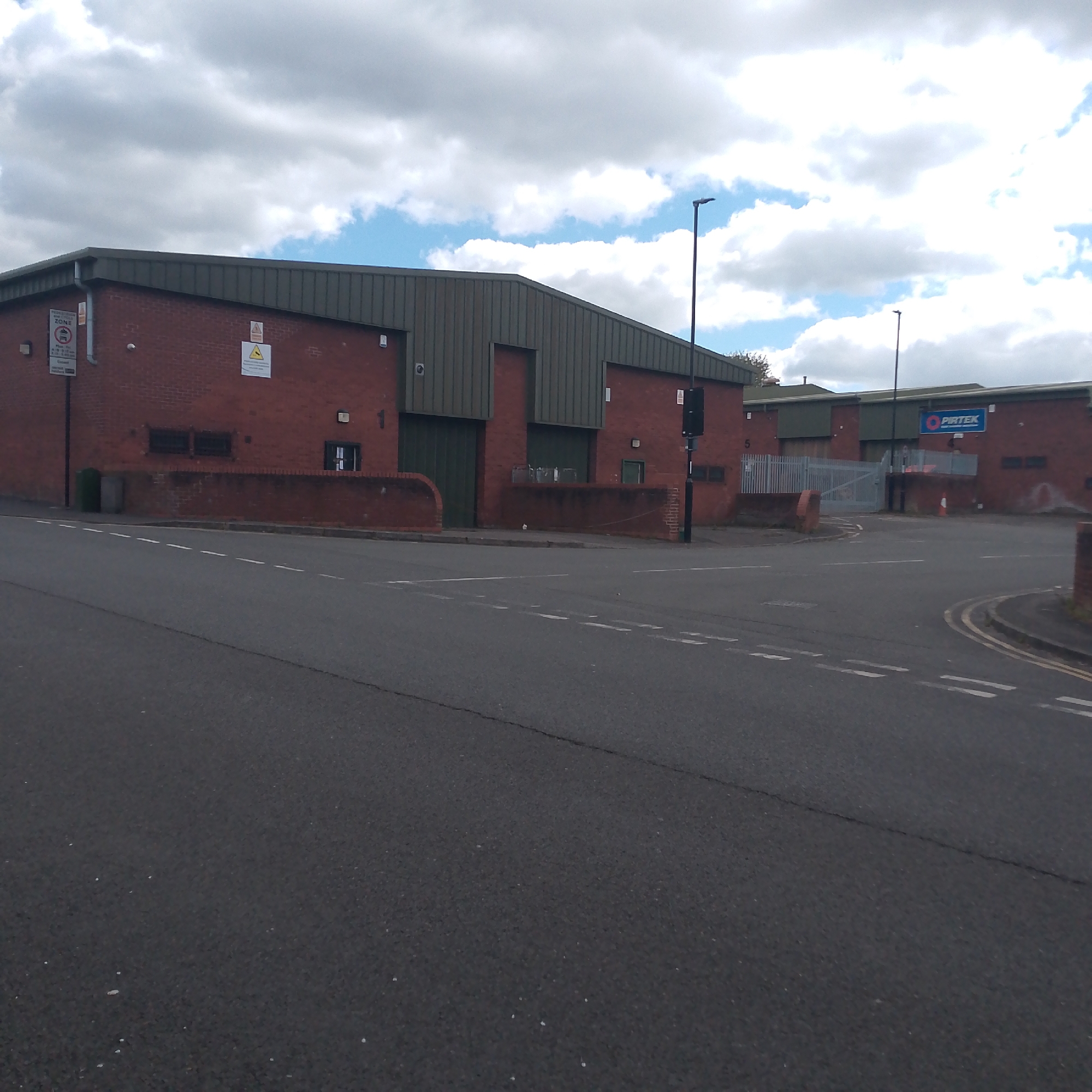 The image depicts a street intersection in an industrial area in Coventry, United Kingdom. The main feature is a brick building with a dark green corrugated metal roof. A sign is visible on the left side of the building, and another warning sign is next to it. There are large green loading bay doors. Adjacent to this building is another similar brick structure with a "PIRTEK" sign. There is a gray metal fence and gate partially visible. Streetlights are present at the roadside. The asphalt road shows painted white lines for traffic guidance. The sky is overcast.