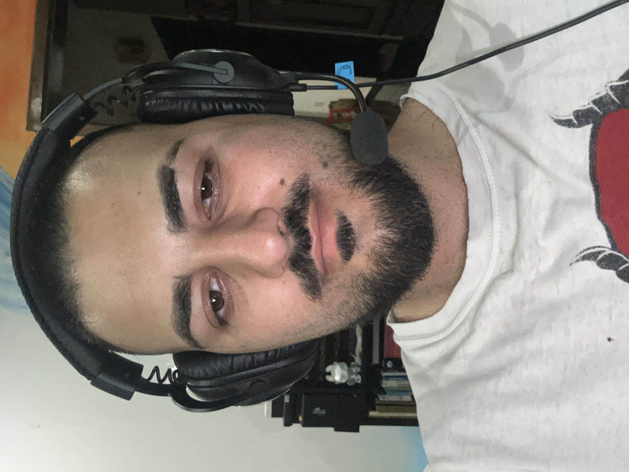A close-up, waist-up selfie of a man wearing black headphones with a microphone boom. He has dark, short hair, a beard, and a mustache. His eyes appear reddish and watery. He is wearing a white t-shirt with a dark graphic design on the front, partially visible. In the background, a bookshelf with various items is visible, along with a light-colored wall and a hint of an orange-toned object. The image appears to be taken indoors, in what could be a home office or gaming setup. There is no visible indication of the specific location in Plovdiv, Bulgaria.