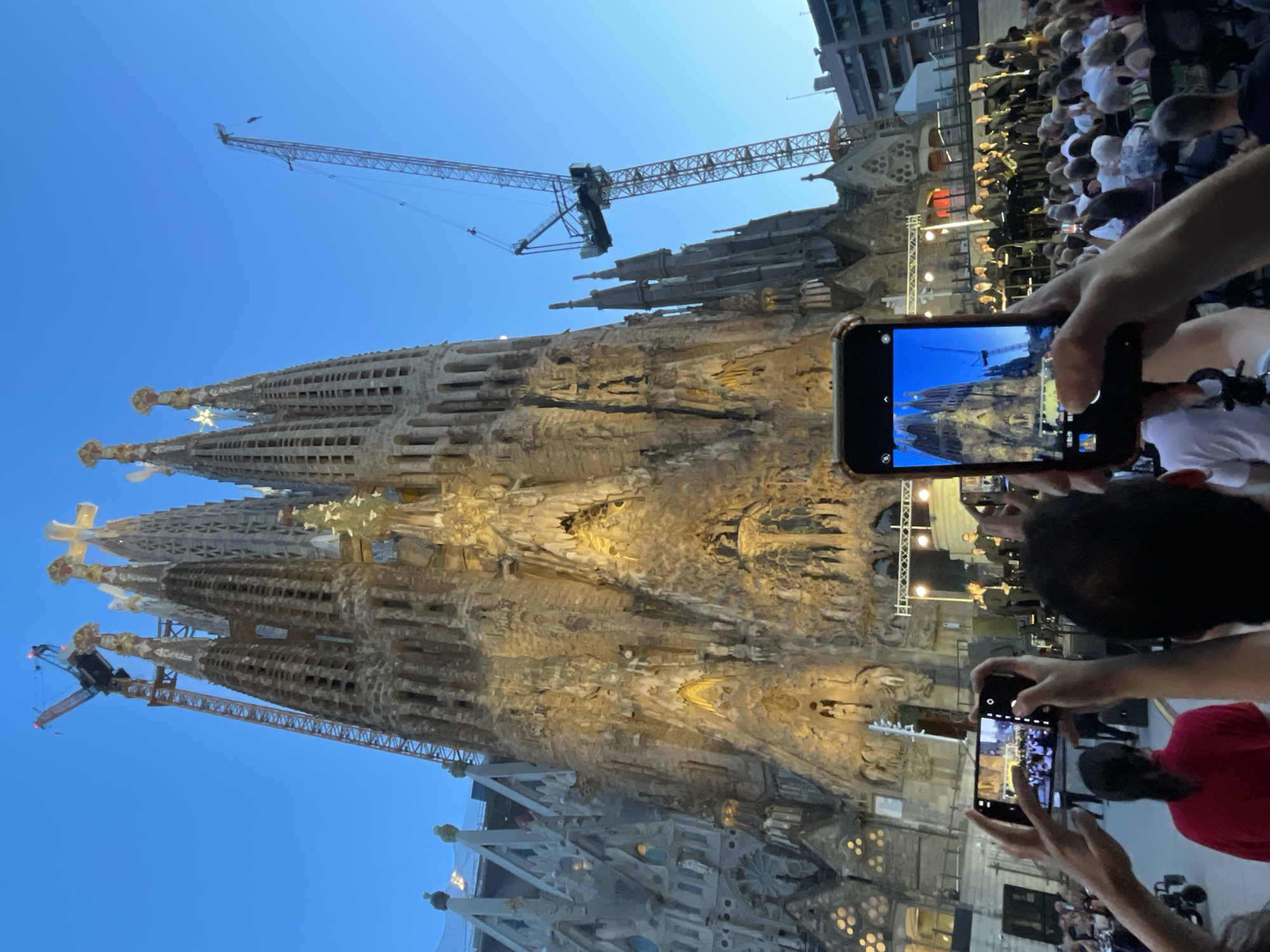 The image captures a crowd gathered in front of the Sagrada Familia in Barcelona, Spain, during an evening event. The iconic basilica, illuminated by warm lights, dominates the background, with construction cranes visible near its spires, indicating ongoing work. In the foreground, several individuals are holding up smartphones to capture the scene, some framing the basilica itself, while others focus on a stage set up with a performing orchestra. The sky is a deep blue, suggesting twilight. The crowd, composed of people of various ages, is seated and standing, attentively watching the performance or taking photographs. The atmosphere appears to be one of a public gathering, possibly a concert or a cultural event, with a blend of appreciation for architecture and the ongoing entertainment.