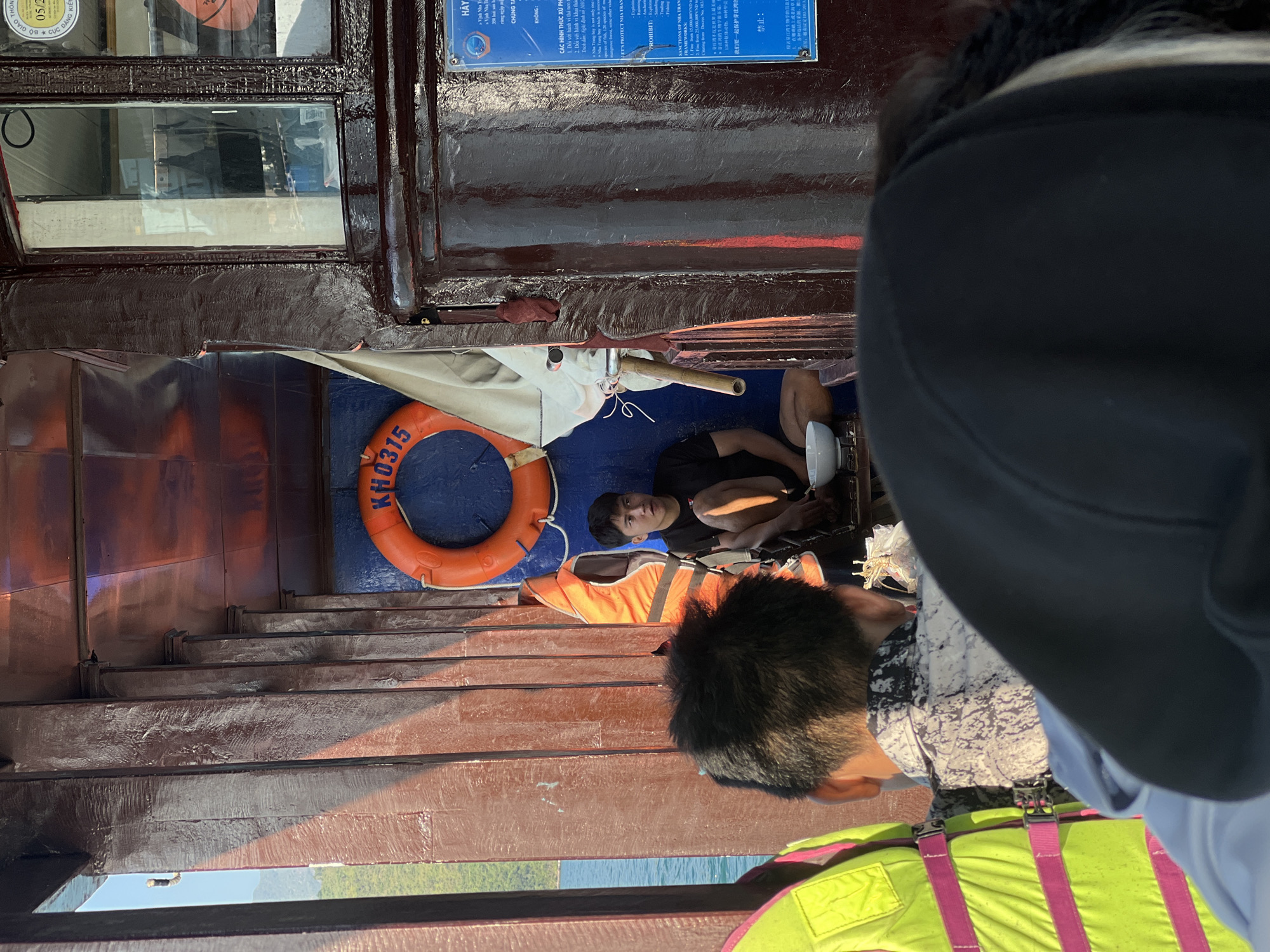 The image captures a scene on a boat, likely in Nam Nha Trang Ward, Vietnam, as inferred from the signs in the frame. A person is sitting on the deck, their appearance is partially obscured by other passengers. Nearby are an orange life buoy marked with "KH0315" and a life jacket. The deck has multiple wooden steps, revealing the boat's structure. A window frame sits atop the scene, with the view outside blurred but suggesting a body of water and greenery. The presence of life-saving equipment suggests a focus on safety in the maritime setting.