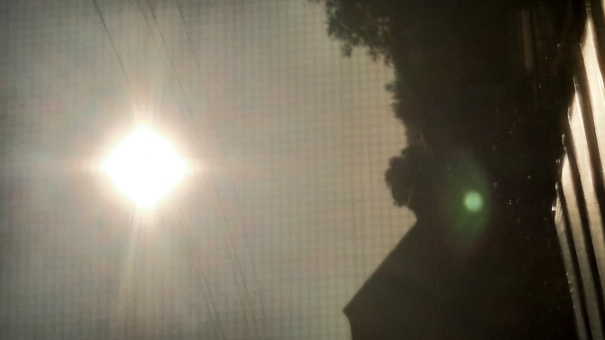 The image shows a silhouette of a house and trees against a bright, square sun. There are power lines visible in the sky. In the foreground, there are horizontal lines, possibly part of a railing or a roof. A green lens flare is present in the lower center of the image. The scene appears to be captured outdoors, possibly from a window or balcony, with the sun high in the sky. The location is identified as Shklov, Belarus. There are no people or animals visible in the image.