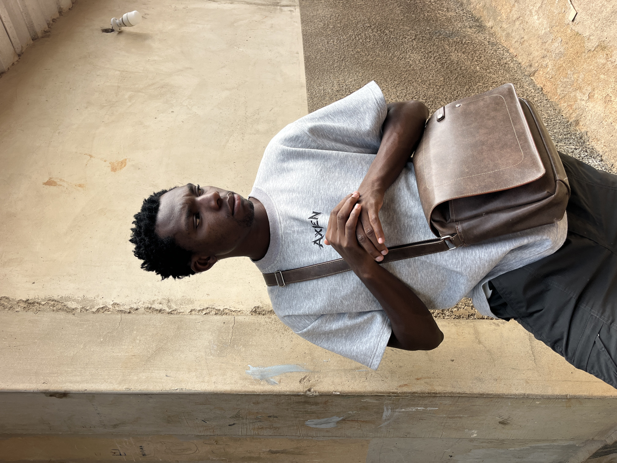 A young Black man is standing outdoors, leaning against a textured, pale yellow wall. He is wearing a light gray sweatshirt with a logo on the chest and dark gray cargo pants. A brown leather messenger bag with a strap is slung across his right shoulder. His hands are clasped in front of him. The background wall has some marks and scuffs, and a small light fixture is visible on the upper right portion of the wall. The ground appears to be dirt or gravel.