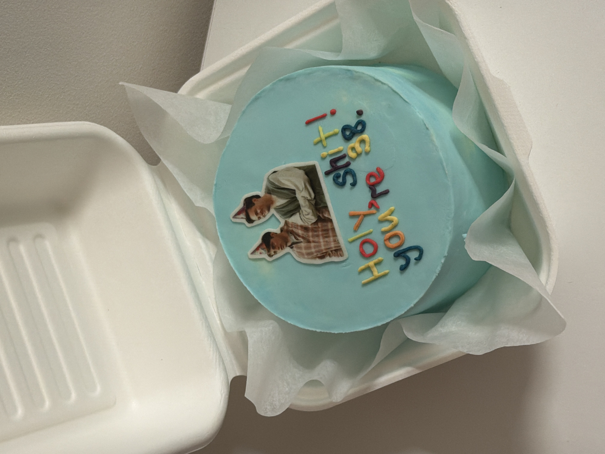 A light blue, round cake is presented inside a white, disposable cake container lined with white tissue paper. The cake features decorative elements, including two identical cut-out images of a man. In the images, the man is shown leaning over, wearing a greyish long-sleeved top, a plaid shirt underneath, and a small red and white party hat.
Edible lettering is piped onto the cake, forming the phrase "HOLY SHIT! you're 38." The text uses a variety of colors: "HOLY" is in orange, "SHIT!" is in red, "you're" is in blue and red, and "38" is prominently displayed in yellow and orange. The cake's design suggests a humorous and informal birthday celebration for someone turning 38.
The cake is positioned centrally within its container, which is slightly open, revealing part of another identical white container to the left. The background is plain and light-colored, indicating an indoor setting, likely with artificial lighting. No specific visual cues indicate the time of day or weather. While the context given is Tel-Aviv, Israel, there are no elements within the image itself to confirm this location.
