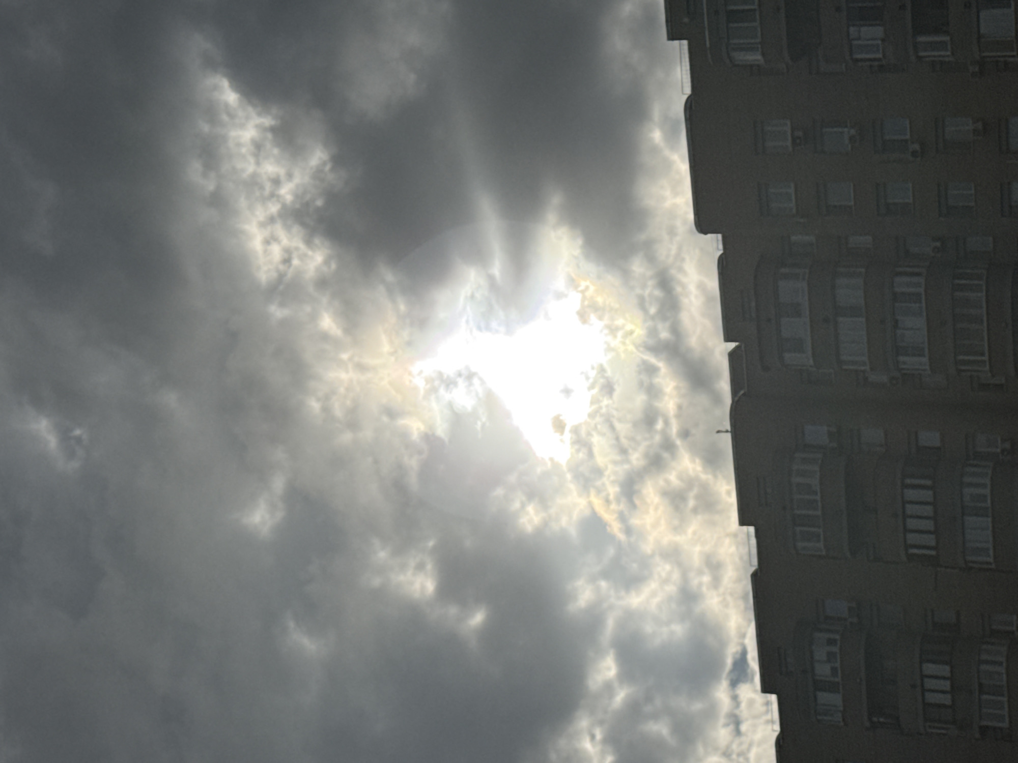 The media file depicts a dramatic sky filled with dark, heavy clouds with the sun breaking through in a radiant burst. The sun's rays are visible, creating a halo effect. The sky is vast and appears to be in motion, suggesting a dynamic weather event, possibly a storm about to break or pass. To the right of the frame, a portion of a tall, multi-story building is visible. The building is constructed of dark material and features numerous windows, some of which appear to have balconies or alcoves. The building's architecture suggests an urban setting. The contrast between the bright, ethereal sky and the solid, geometric form of the building creates a striking visual. The time of day is not explicitly indicated, but the strong sunlight suggests it could be daytime. The weather is clearly overcast with a significant amount of cloud cover, though the sun's presence indicates it is not entirely dark. The overall mood is one of power and drama, with the natural forces of the sky dominating the scene. There is no visible text in the media file. The location context is Moscow, Russia.
