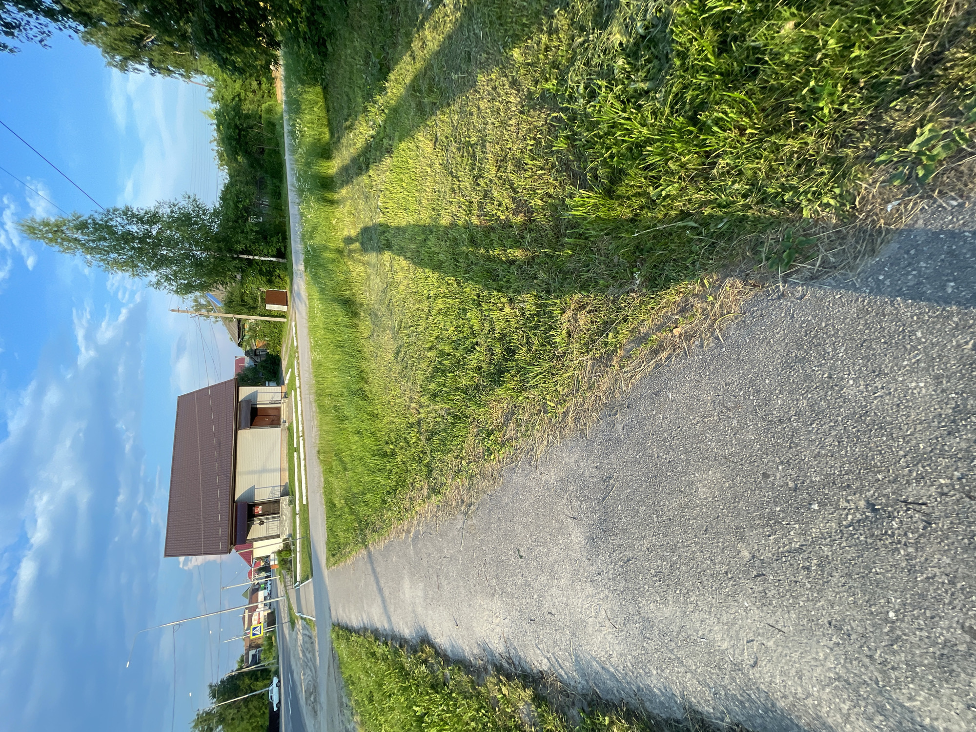 This media file displays a wide-angle, vertically oriented view of a sidewalk adjacent to a grassy area and a residential street. The scene appears to be a rural or suburban setting in Красные Баки, Russia. The time of day is likely late afternoon or early evening, given the long shadows cast by trees and an unidentified object, which is likely the photographer.
The subjects include a paved sidewalk with a strip of grass bordering it on one side and a street on the other. The street has a few parked cars and houses or buildings visible in the background. One prominent building has a brown, pitched roof and light-colored walls, with a small porch area. Power lines and streetlights are also visible along the street.
The weather seems pleasant, with a clear blue sky dotted with white clouds. The sun is shining, illuminating the green grass and casting distinct shadows. The overall emotion conveyed is one of calm and everyday life in a peaceful setting.
Notable details include the shadow of a person on the grassy area, suggesting the image was taken by someone walking or standing there. The perspective is unusual, giving a sense of being close to the ground and looking upwards at the sky and surroundings. There is no visible text in the image.