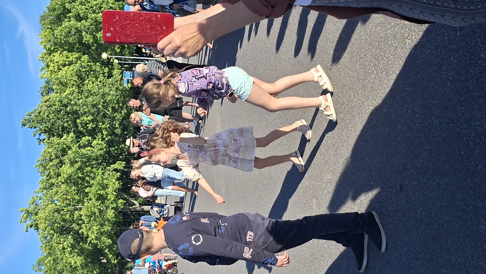 In Tallinn, Estonia, on a sunny day with a clear blue sky, a group of people are gathered outdoors. The ground is paved, suggesting a public area like a street or plaza, with large trees lining the background. In the foreground, a young boy wearing a dark long-sleeved shirt, dark pants, and a baseball cap is walking away from the camera. To his right, two young girls are dancing. The girl on the left wears a light-colored, short-sleeved dress with a floral pattern and a pink headband. The girl on the right is dressed in a purple long-sleeved top with a graphic design and light blue shorts. Both girls are wearing sandals. Behind them, a crowd of adults are visible, some standing and observing, while others appear to be engaged in conversation. One person in the upper right corner is holding up a red phone, as if taking a photo or video. The scene suggests a casual outdoor event or gathering.