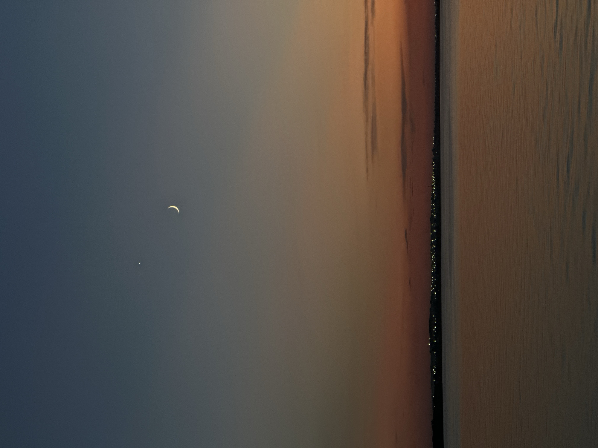 This image captures a serene twilight scene over a body of water, likely a lake or sea, in Kokshetau, Kazakhstan. The sky transitions from a deep blue on the left to a warm orange and pink gradient on the right, indicative of either sunrise or sunset. A slender crescent moon is visible in the dusky blue portion of the sky, accompanied by a bright star or planet.
The right side of the image reveals a distant shoreline dotted with numerous small, twinkling lights, suggesting a town or city settlement. These lights create a subtle, shimmering line against the darker landmass. The water itself is calm, reflecting the colors of the sky with a soft, textured surface. The overall mood is peaceful and tranquil, evoking the quiet beauty of the transitional hours between day and night. No people or distinct objects are discernible, focusing the viewer's attention on the natural elements of the sky and water, and the man-made lights on the horizon. The image does not contain any visible text.
