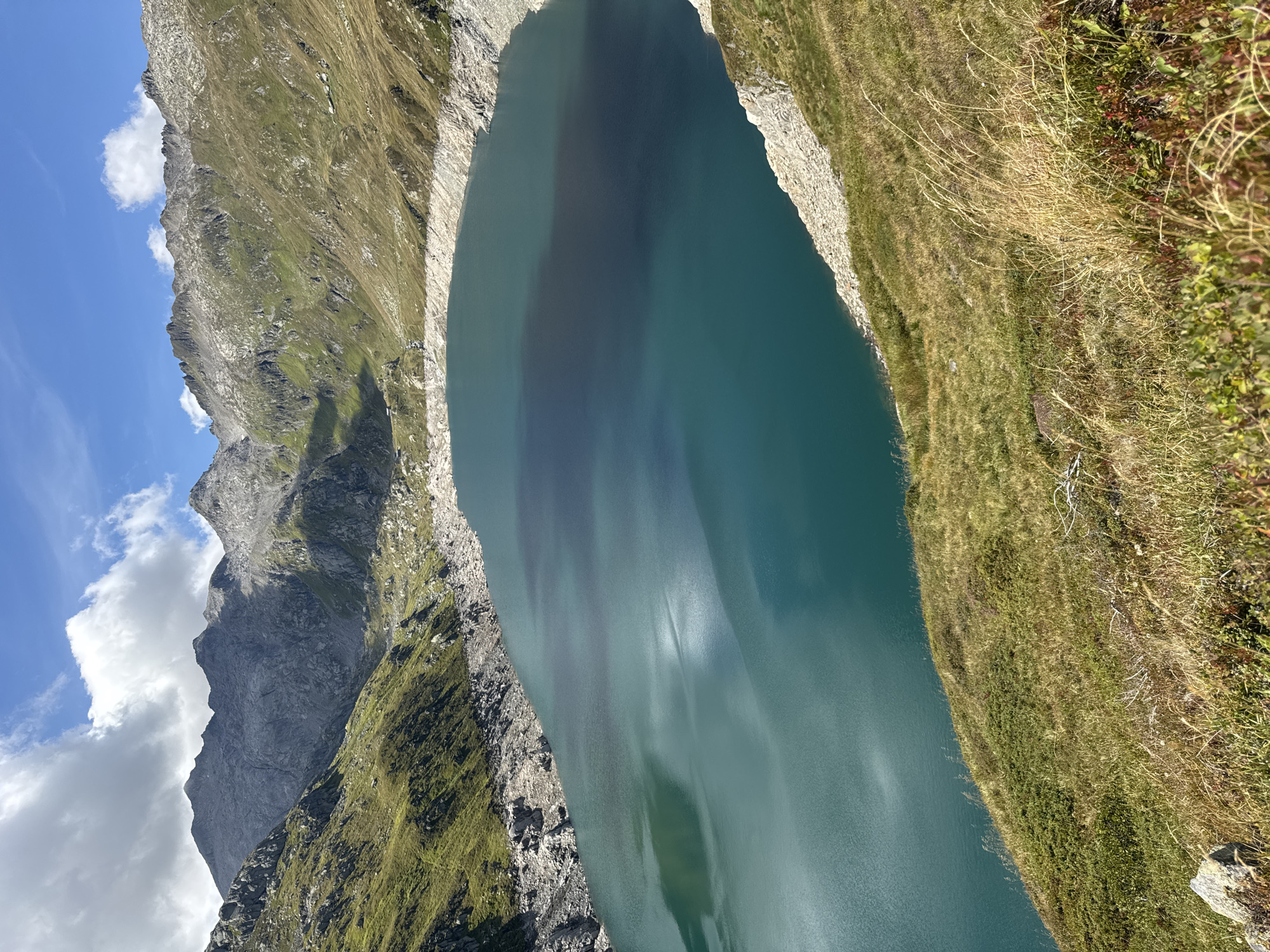 This is a high-angle, landscape photograph of a large, tranquil lake surrounded by steep, rocky, and vegetated mountain slopes under a partly cloudy blue sky. The lake's water is a deep teal color, with lighter reflections and ripples visible on its surface. The shoreline is primarily composed of exposed rock and scree. The mountains rise dramatically from the lake, with patches of green grass and darker vegetation clinging to their inclines. The sky above features scattered white clouds against a clear blue expanse. The foreground consists of a grassy slope with some dried grasses and a few rocks. This scene is consistent with the landscape around the artificial reservoir of Lago della Nose, located near Airolo, Switzerland, which is known for its dramatic alpine scenery. There are no people or animals visible in the image.