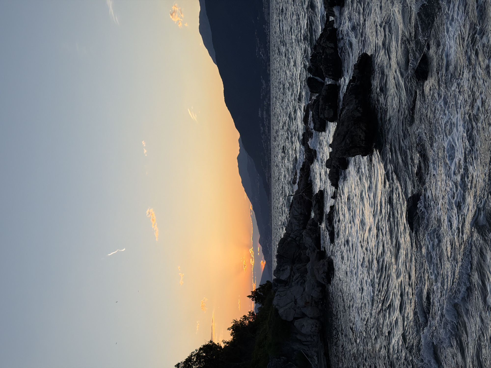 The scene depicts the coastal landscape of Gošići, Montenegro, at sunset. The sky transitions from pale blue to orange as the sun dips behind a dark mountain range on the horizon. The sea is choppy with white-capped waves reflecting the fading light. Rocky outcrops line the shore, their dark forms contrasting with the sea's silvery surface. The silhouette of trees and vegetation marks the shoreline. Three small birds are visible in the sky.