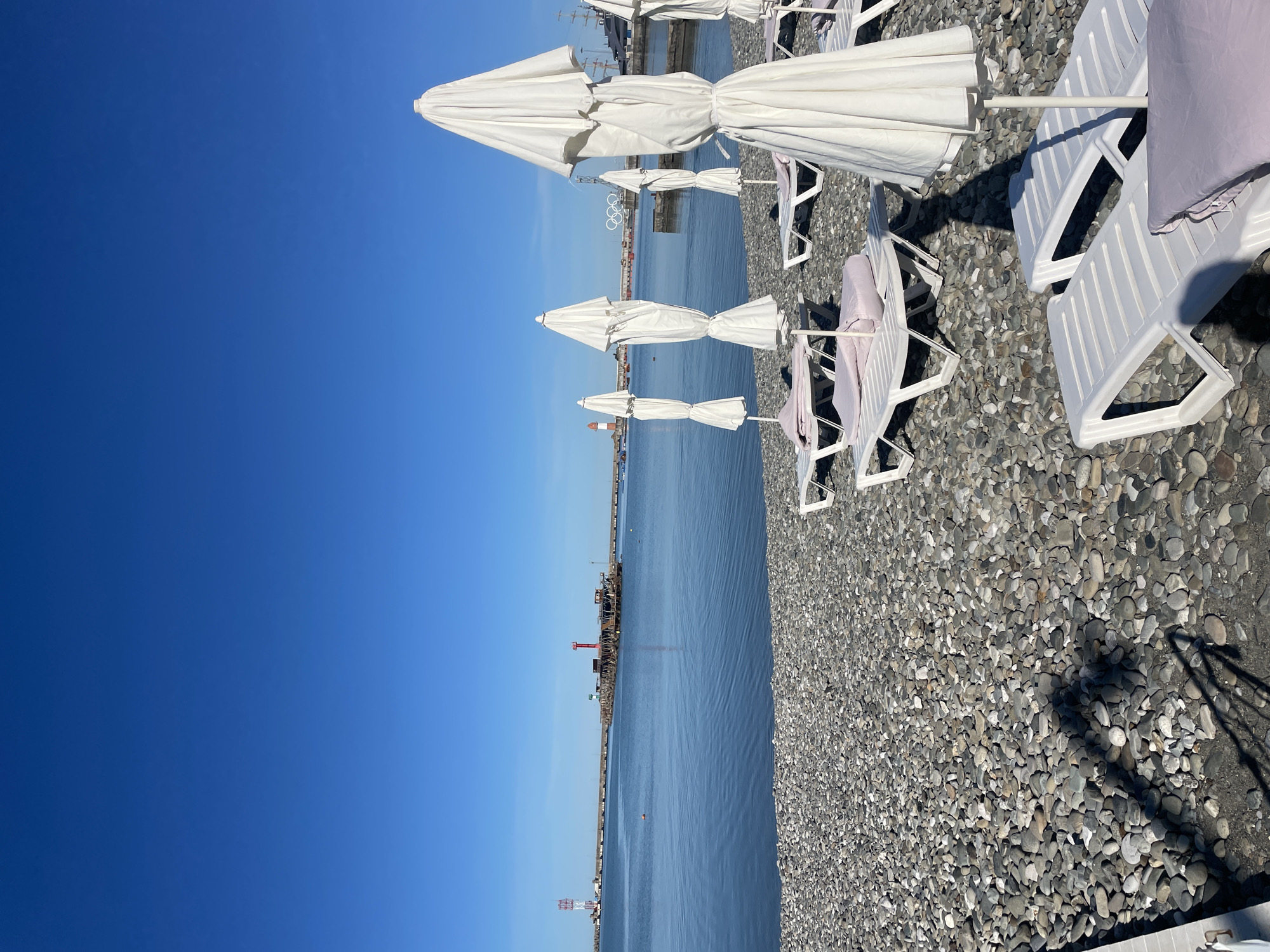 The image captures a peaceful, sunny day at a pebble beach in Sochi, Russia. The scene is oriented as if looking out from the shore towards a vast expanse of calm, deep blue water, likely the Black Sea.
In the foreground and mid-ground, the beach is covered in smooth, rounded pebbles, predominantly in shades of grey and white. A neat row of white, closed beach umbrellas stands upright along the water's edge, casting long, sharp shadows onto the stony ground, indicating bright sunlight. Behind these umbrellas, several white plastic lounge chairs are arranged, some fitted with light purple cushions or towels, suggesting a ready but currently unoccupied relaxation area.
The water is remarkably still, reflecting the brilliant blue of the sky with only minor ripples disturbing its surface. Along the distant horizon, various structures are visible. These include what appear to be port facilities with cranes, buildings, and possibly a lighthouse or tall antenna. A significant landmark, the white structure of the Olympic rings, can be clearly identified across the water, providing a strong contextual clue to the location as Sochi, the host city of the 2014 Winter Olympics. A few small buoys or markers are scattered in the water in the distance.
The sky above is a uniform, vivid blue, completely free of clouds, signifying clear weather. The bright sunlight and distinct shadows suggest it is either morning or late afternoon. No people are visible on the beach or in the water, contributing to an atmosphere of quiet serenity and readiness for activity. There is no visible text in the image.
