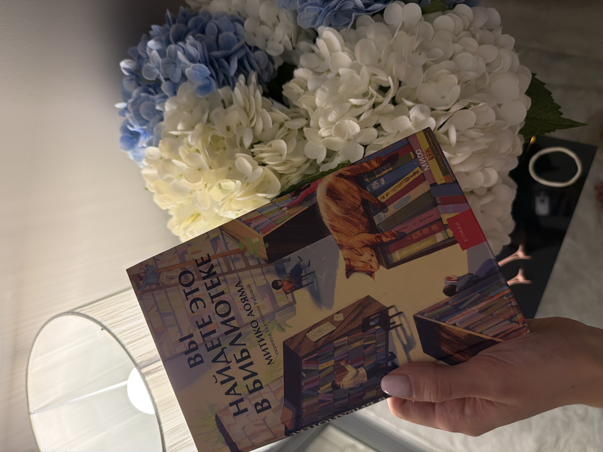 A person holds a book titled "Вы Найдете Это в Библиотеке" (You'll Find It in the Library) by Mitiko Aoyama. The book cover depicts a library scene with a cat. In the background, there's a bouquet of white and blue hydrangeas. A lamp is visible in the lower left corner. The image was likely taken indoors, based on the lighting and objects present. The location in the city of Khimki, Russia is not possible to verify based on only the provided image.