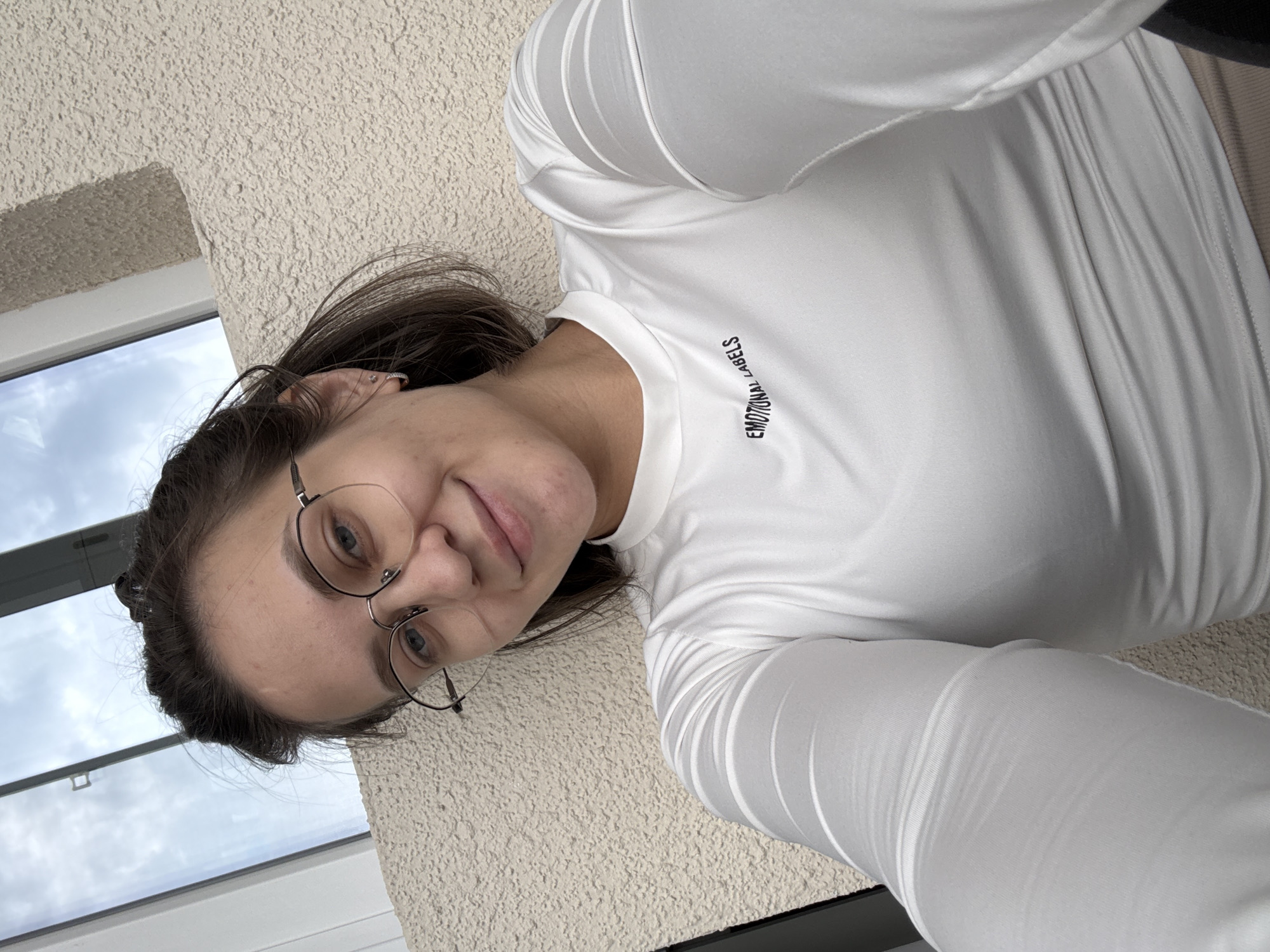 A young woman is depicted in a selfie, taken from a slightly high angle and rotated counter-clockwise. She has dark hair, pulled back, and wears round, thin-framed glasses. A gentle smile plays on her lips, and her eyes, which appear blue, gaze upwards towards the camera.
She is wearing a form-fitting, long-sleeved white top, possibly athletic wear, which features subtle ribbed patterns on the sleeves and body. Across the chest area of her top, the words "EMOTIONAL LABELS" are printed in black, uppercase letters. The crops suggest she might be wearing matching light-colored bottoms, possibly leggings, completing an activewear ensemble. A small, shiny ear piercing is visible in her right earlobe.
The background consists of a textured, light-colored wall, likely stucco or rough plaster, visible to her right and below her. To her left, a window shows a cloudy, overcast sky, indicating daytime with diffused light. The window frame appears light-colored. The lighting is soft and even, highlighting her features without harsh shadows. Her expression conveys a sense of calm and contentment.