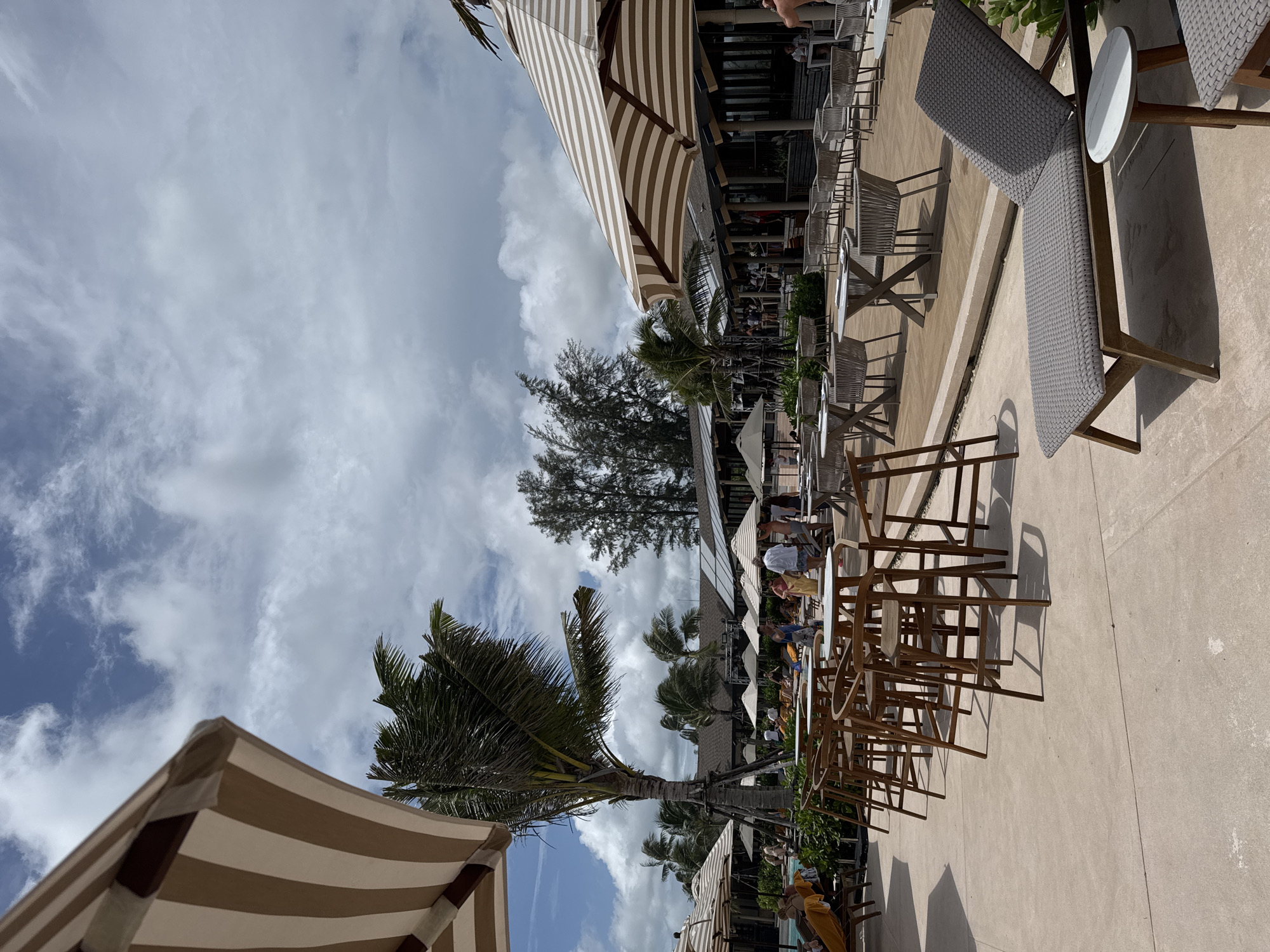 The image depicts an outdoor scene in Choeng Thale, Thailand, featuring a patio or dining area with tables and chairs, umbrellas, and palm trees. People are seated at some of the tables. The sky is cloudy.