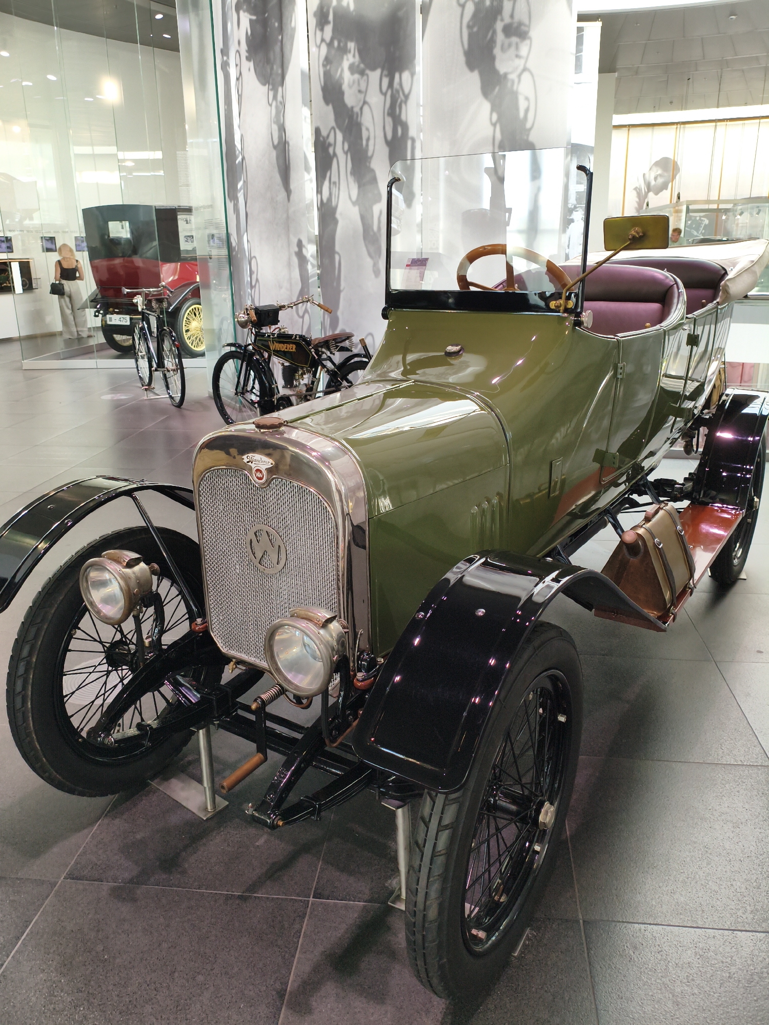 A green vintage touring car is displayed in a museum, likely the Audi Museum in Ingolstadt, Germany. The car is positioned at a slight angle, facing towards the right of the frame, with its front end closest to the viewer. It features a chrome grille with a circular emblem, two prominent headlights, and a wooden steering wheel visible through the windshield. The car has burgundy upholstered seats and a folded tan fabric roof. Attached to the side of the car is a brown leather-bound storage box. Behind the touring car, a black vintage motorcycle with the name "Wanderer" is visible. Further in the background, a red and black vintage automobile with yellow wheels is also on display, alongside a woman wearing beige trousers and a black top, looking at the red car. The environment appears to be a museum with polished tile flooring and large glass windows, revealing abstract black and white wall art in the background.