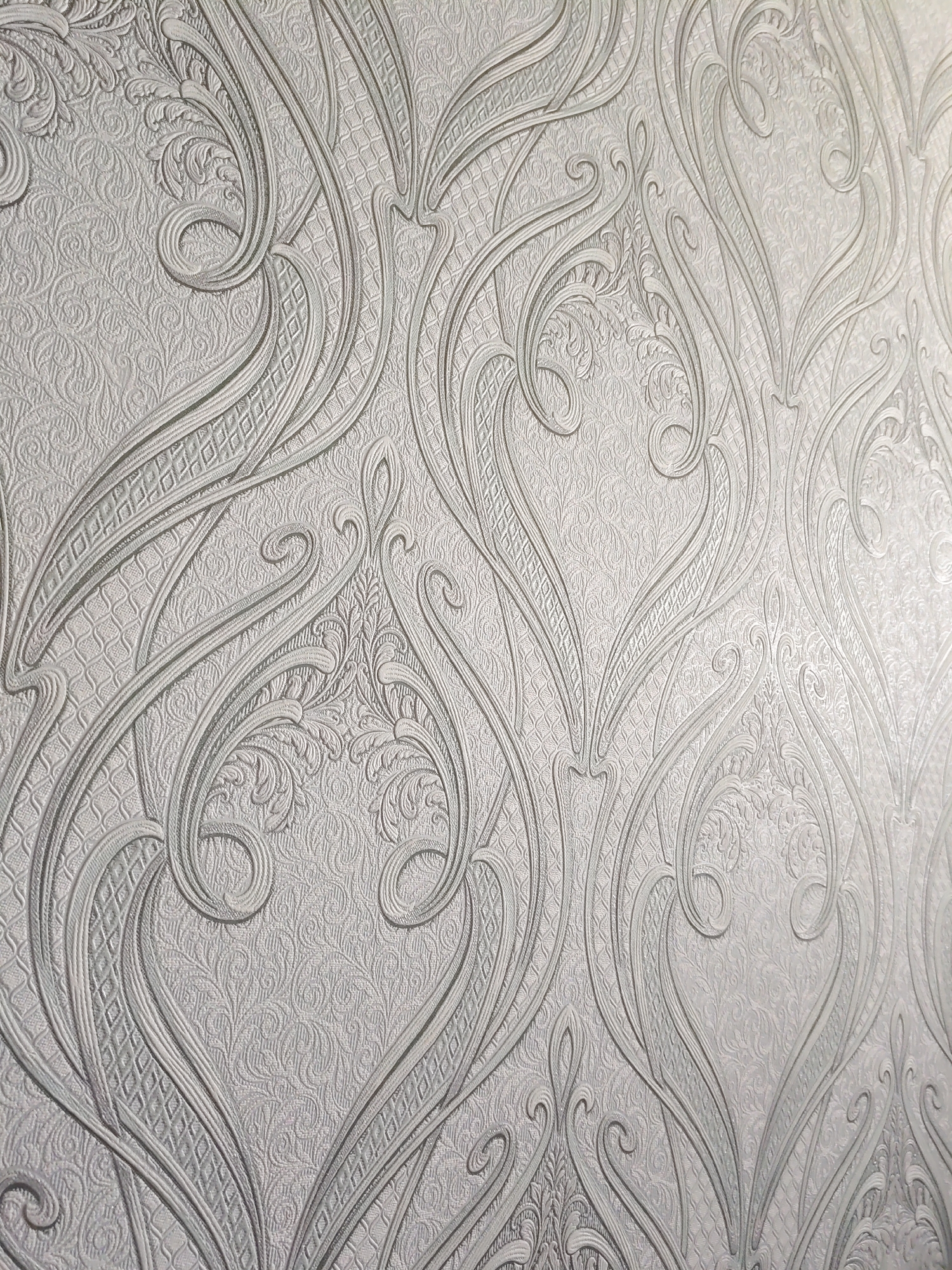 The image displays a close-up view of a textured wallpaper with an ornate, damask-like pattern. The design features raised, scrolling floral motifs and swirling lines in a monochromatic silver or light gray color. The background of the wallpaper has a subtle, embossed texture, possibly resembling small, repeating floral or geometric shapes. The lighting highlights the three-dimensional quality of the raised patterns, creating shadows that accentuate the depth and detail of the design. There are no people or any other objects visible in the image, and the setting appears to be an interior space. The location in the city of Курашим, Russia is not determinable from the image itself.