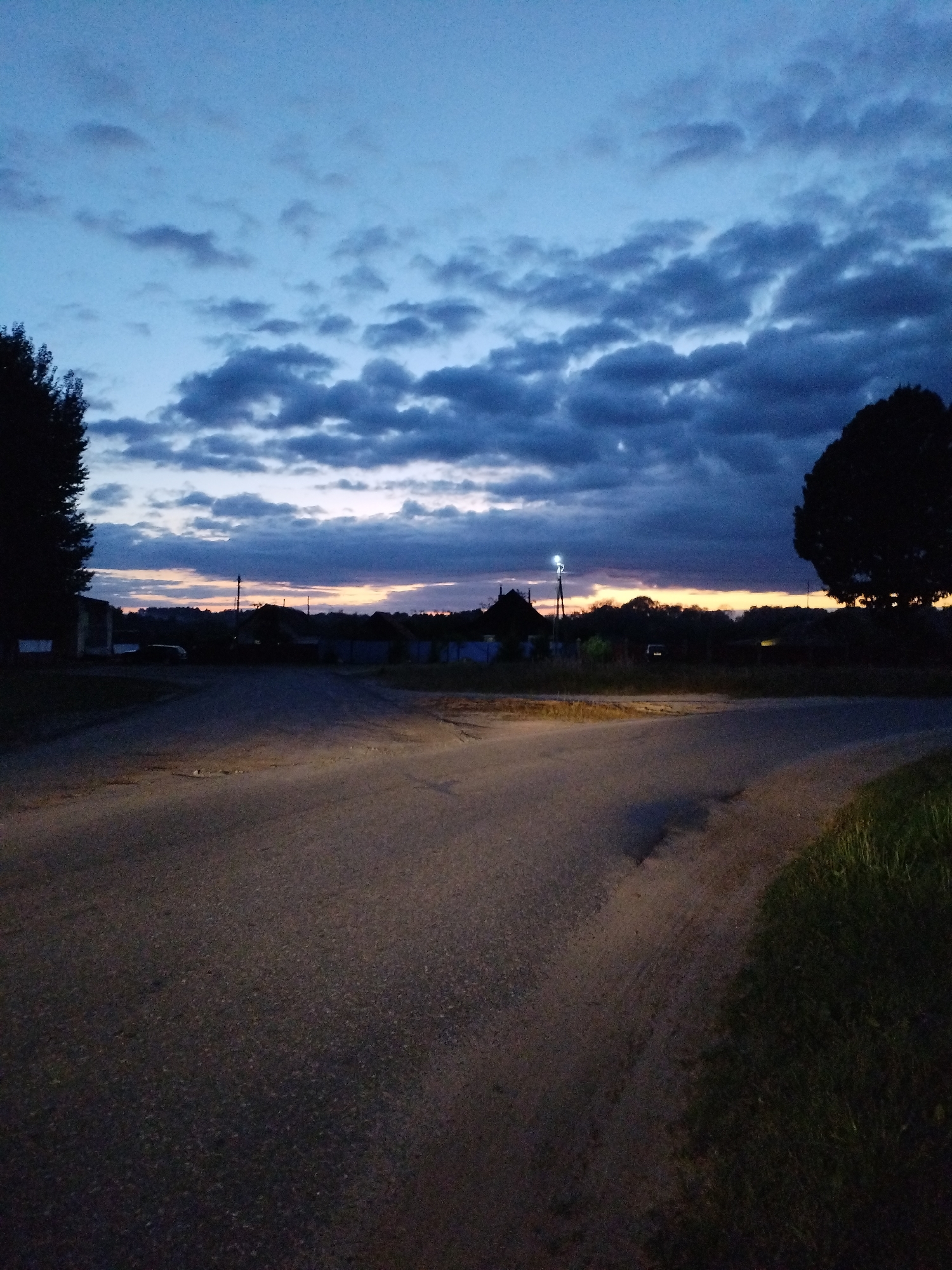 The image depicts a road intersection at dusk in Bialynichy, Belarus. The sky is a deep blue with scattered, dark clouds. A sliver of orange light is visible on the horizon. Silhouettes of trees and houses are present on either side of the road, along with a few faint lights from buildings. The road surface is asphalt, with some visible patches and texture. There are no people or vehicles clearly visible in the scene. The overall impression is a quiet, rural evening setting.