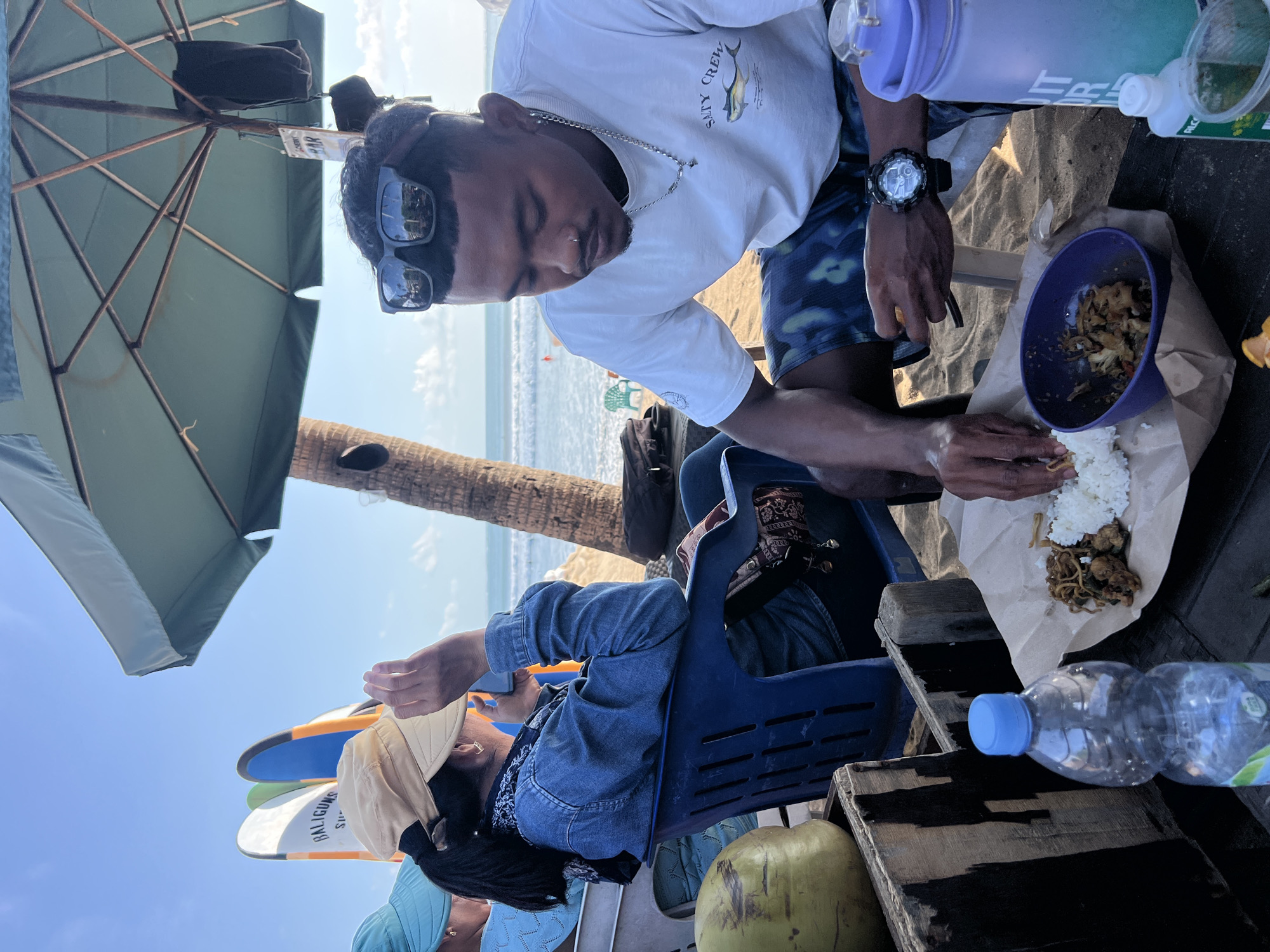 In Kuta, Indonesia, on a sandy beach, two individuals are seated outdoors under a large umbrella. In the foreground, a man wearing sunglasses, a white t-shirt with a "Salty Crew" graphic, and patterned shorts is eating rice and noodles from a plate. He is also wearing a watch. To his left, a person wearing a denim jacket and a light-colored hat is holding a phone to their ear, seated in a blue plastic chair. In the background, surfboards are visible, and the ocean with waves is in the distance. A coconut and a plastic water bottle are on a wooden surface in the left of the frame. A large purple water bottle is on the right. The scene suggests a casual meal or break on the beach.