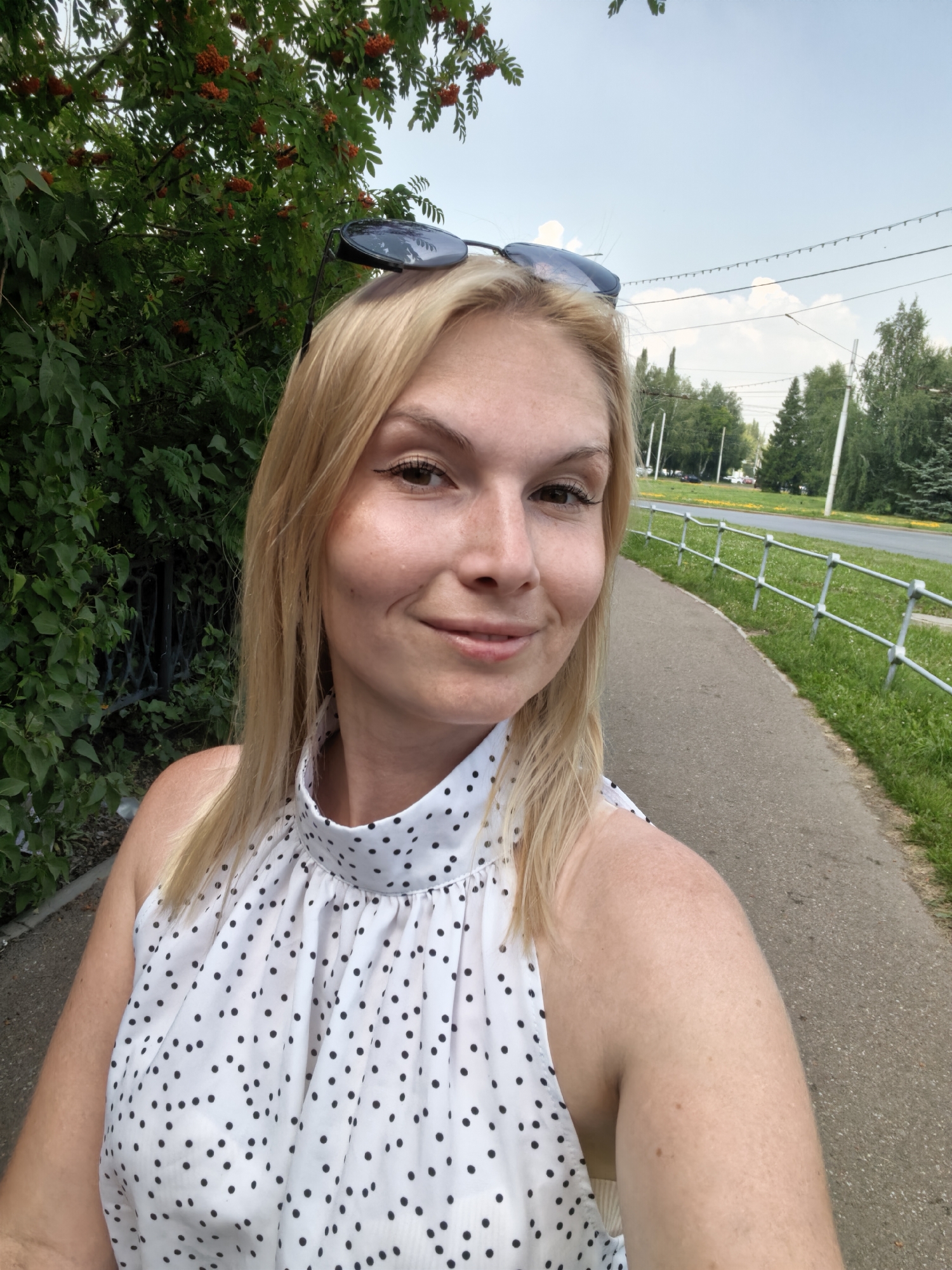 A blonde woman with sunglasses perched on her head takes a selfie outdoors. She is wearing a white sleeveless top with black polka dots. Behind her, lush green foliage with red berries is visible on the left, and a sidewalk and road bordered by a metal fence are on the right. The sky is partly cloudy. The scene appears to be in a park or along a street in Sterlitamak, Russia, given the presence of deciduous trees and the overall urban park-like setting.