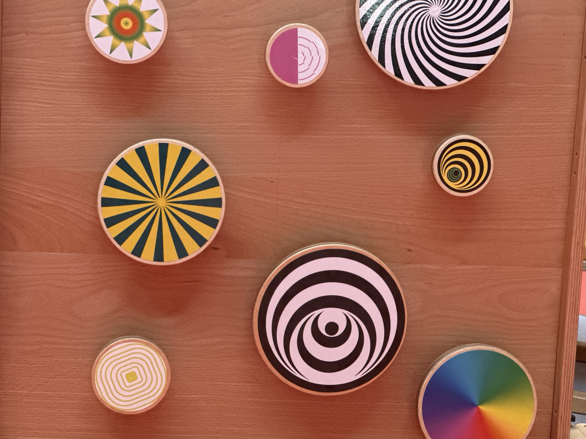 The image displays a collection of circular, spinning toys with optical illusion patterns arranged on a wooden surface. There are eight such toys visible in the frame.
Starting from the top left and moving clockwise, the first toy features a starburst pattern with green and orange colors radiating from a central red circle. To its right, a smaller toy has a pinkish-purple half-circle pattern with concentric white lines, resembling an onion slice. Further to the right, a larger toy exhibits a black and white spiral pattern, creating a strong dizzying effect. Below this, a very small toy displays concentric yellow and black circles with a green center.
Moving to the bottom row, a toy on the left shows a square spiral pattern in yellow and white. To its right, a large circular toy has alternating black and white stripes that form a concentric, inward-moving pattern, resembling a stylized eye or a vortex. Below the black and white spiral toy, to the right, is a circular toy with a rainbow color gradient, with segments of red, orange, yellow, green, blue, and purple radiating from the center. Finally, to the left of the rainbow toy and below the yellow and white spiral toy, is a large circular toy with a radiating pattern of dark teal and bright yellow stripes.
The toys are mounted on a light brown wooden surface, which appears to be a wall or a board, possibly in a playroom or educational setting given the nature of the toys. The lighting is even and suggests an indoor environment. There is no visible text in the image. The overall impression is one of a playful and visually engaging display.