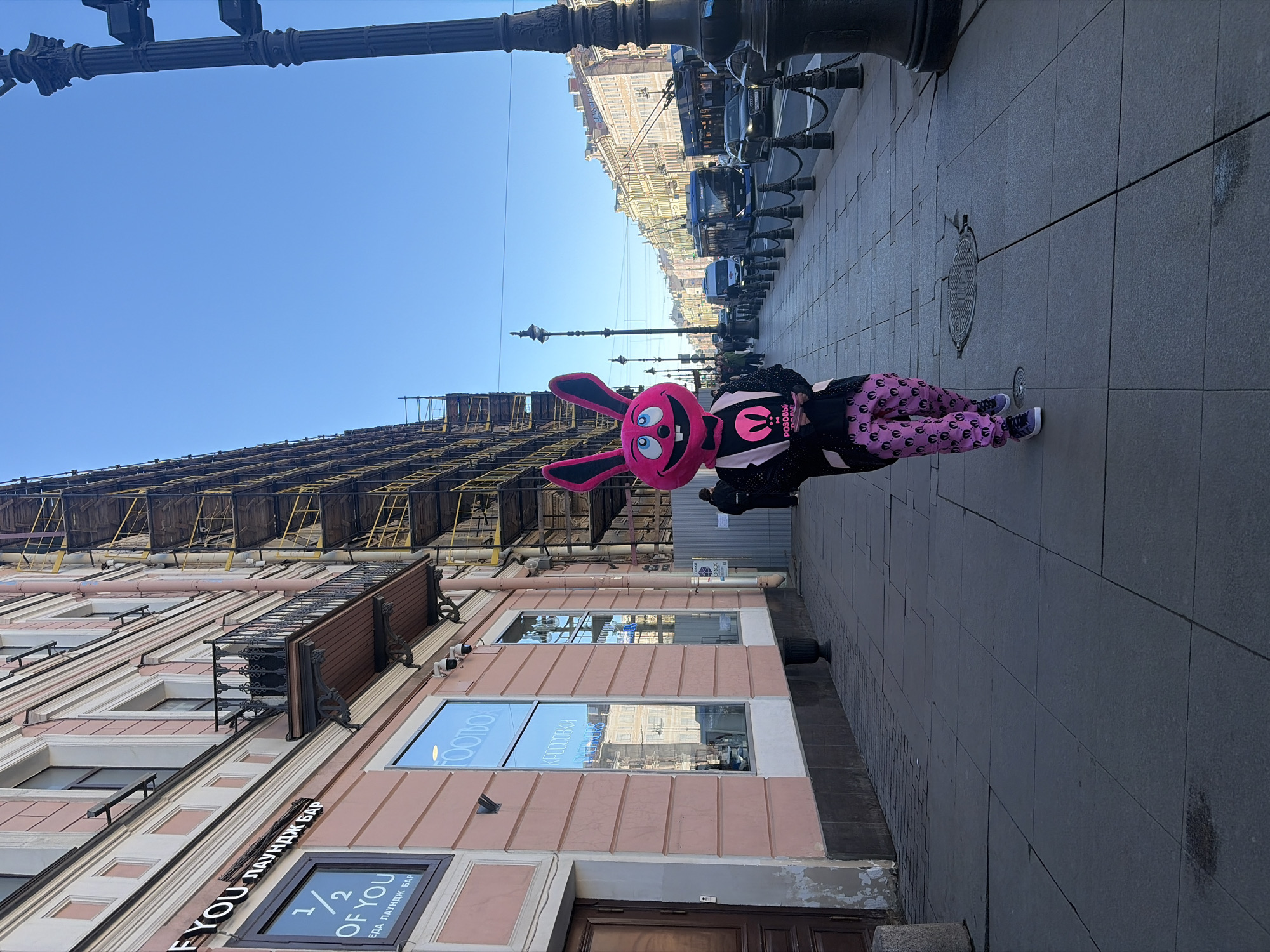 The image shows a person wearing a large pink rabbit costume standing on a sidewalk in Saint Petersburg, Russia. The person is dressed in pink pants with cartoon rabbit face patterns and a black jacket with a logo featuring a stylized bunny. The building next to the person has a sign that reads "1/2 OF YOU". Scaffolding covers the upper part of the building. A line of dark-colored vehicles are parked along the street behind the person. The sky is clear and blue.