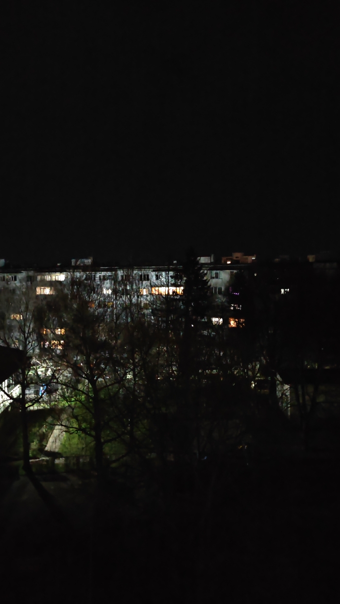 The image displays a nocturnal view of an urban residential area in Shumen, Bulgaria. In the background, a multi-story residential building with a light-colored facade is visible, featuring multiple windows, many of which are illuminated with warm, yellow-orange light from interior sources. The sky above the building is uniformly dark. The midground and foreground are heavily obscured by the dark silhouettes of numerous bare deciduous trees and some patches of dimly lit green foliage, suggesting an unseen light source to the left. A triangular architectural element is partially discernible within the lower left foreground, obscured by vegetation. No individuals or specific activities are discernible within the frame.