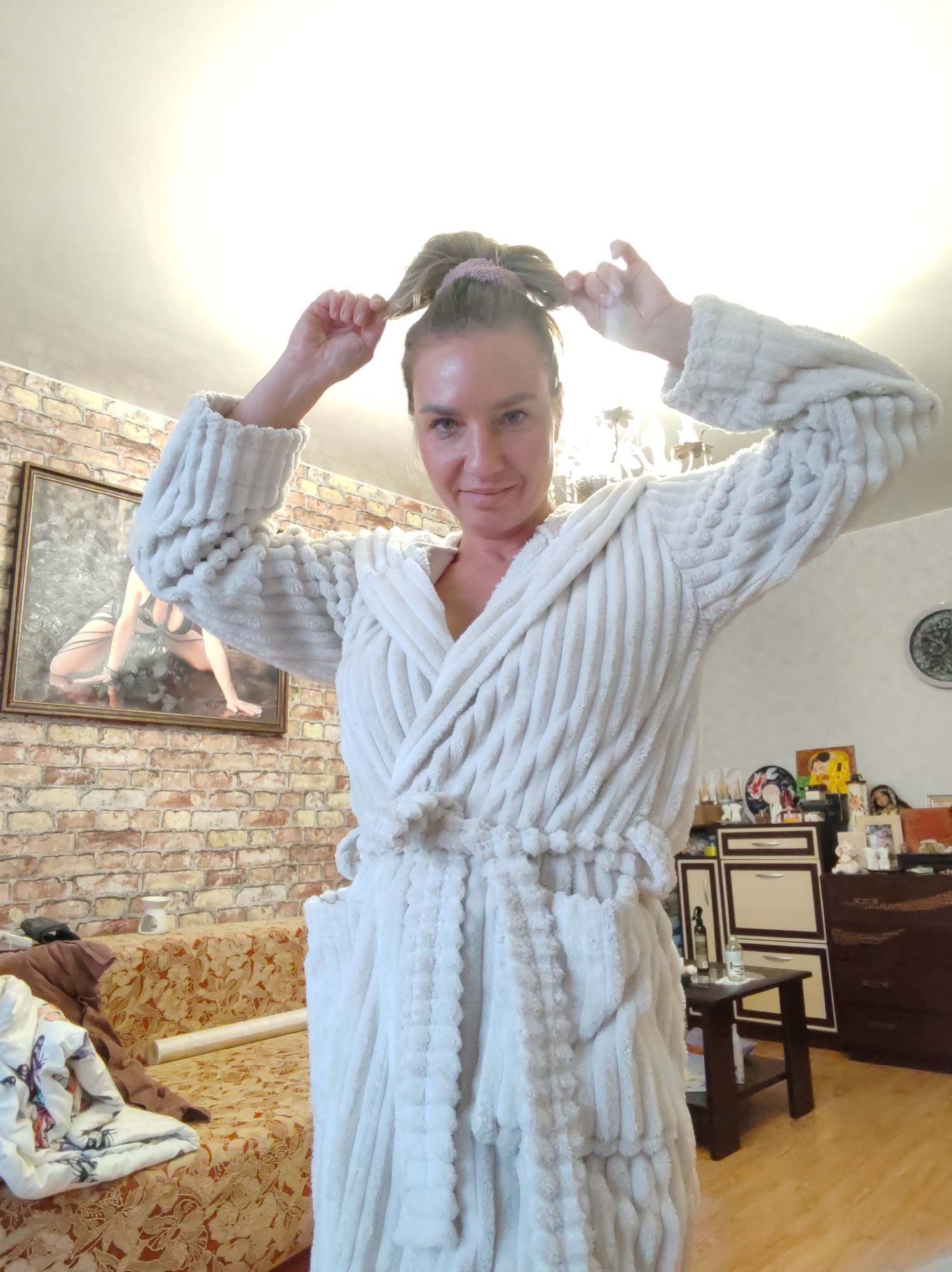 A woman in a white bathrobe stands indoors, holding her hair up with both hands. Behind her is a room with a brick wall, artwork, and furniture including a couch and cabinets. The location is in a residential setting, presumably in Petropavlovsk-Kamchatsky, Russia.