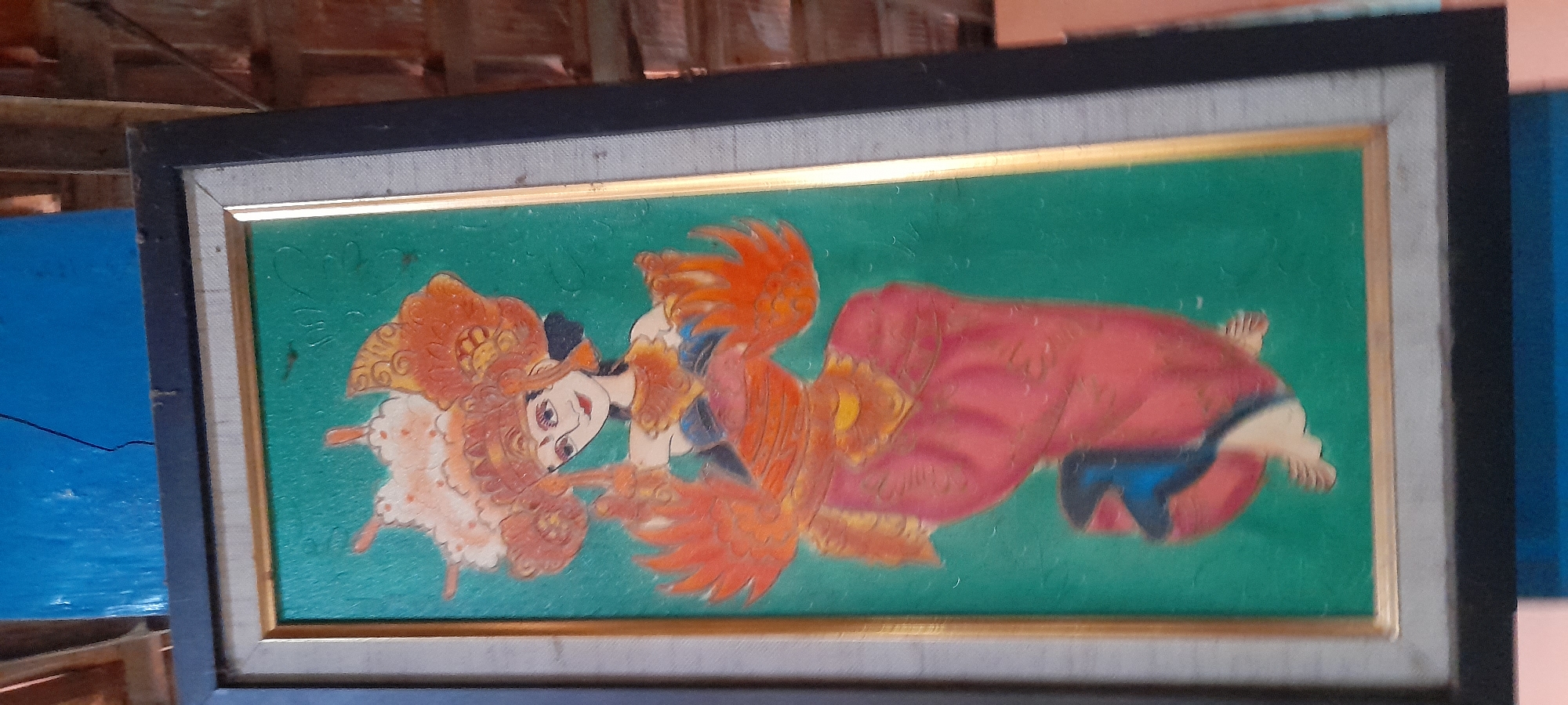 A framed painting depicts a Balinese dancer with elaborate headdress and vibrant orange and red attire against a green background. The dancer is in a graceful pose, with hands extended and adorned with feathered adornments. The painting is framed with a dark outer frame and a gold inner frame. The artwork is displayed indoors, with a blurred background showing architectural elements, possibly a wall painted blue and a brick structure. The overall lighting is dim.