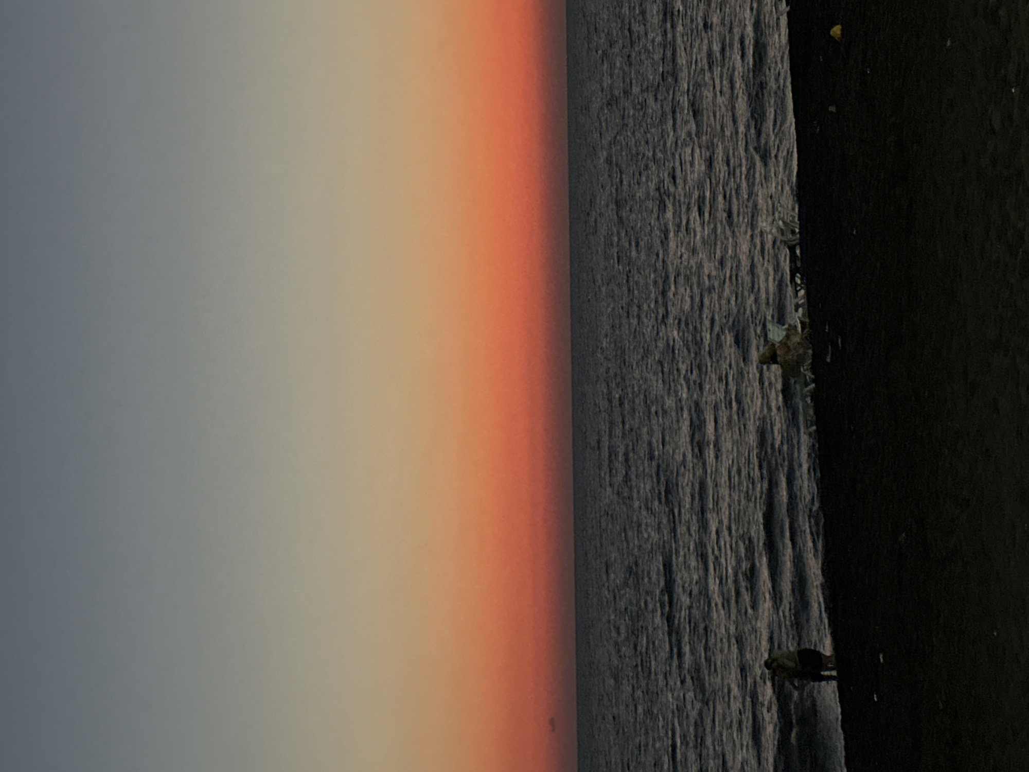 A vertical, low-angle, wide shot captures a twilight beach scene. The sky is a gradient of soft blue at the top, transitioning through pale yellow and into a vibrant orange-red sunset band just above the horizon. The ocean below is a dark, undulating expanse, reflecting the colors of the sky. The foreground is a dark, sandy beach. On the left side of the beach, two figures stand near the water's edge, appearing to be interacting. To the right, several indistinct shapes suggest debris or objects scattered on the sand. A small yellow object is visible further to the right on the beach. The overall impression is one of a serene and colorful evening at the coast.