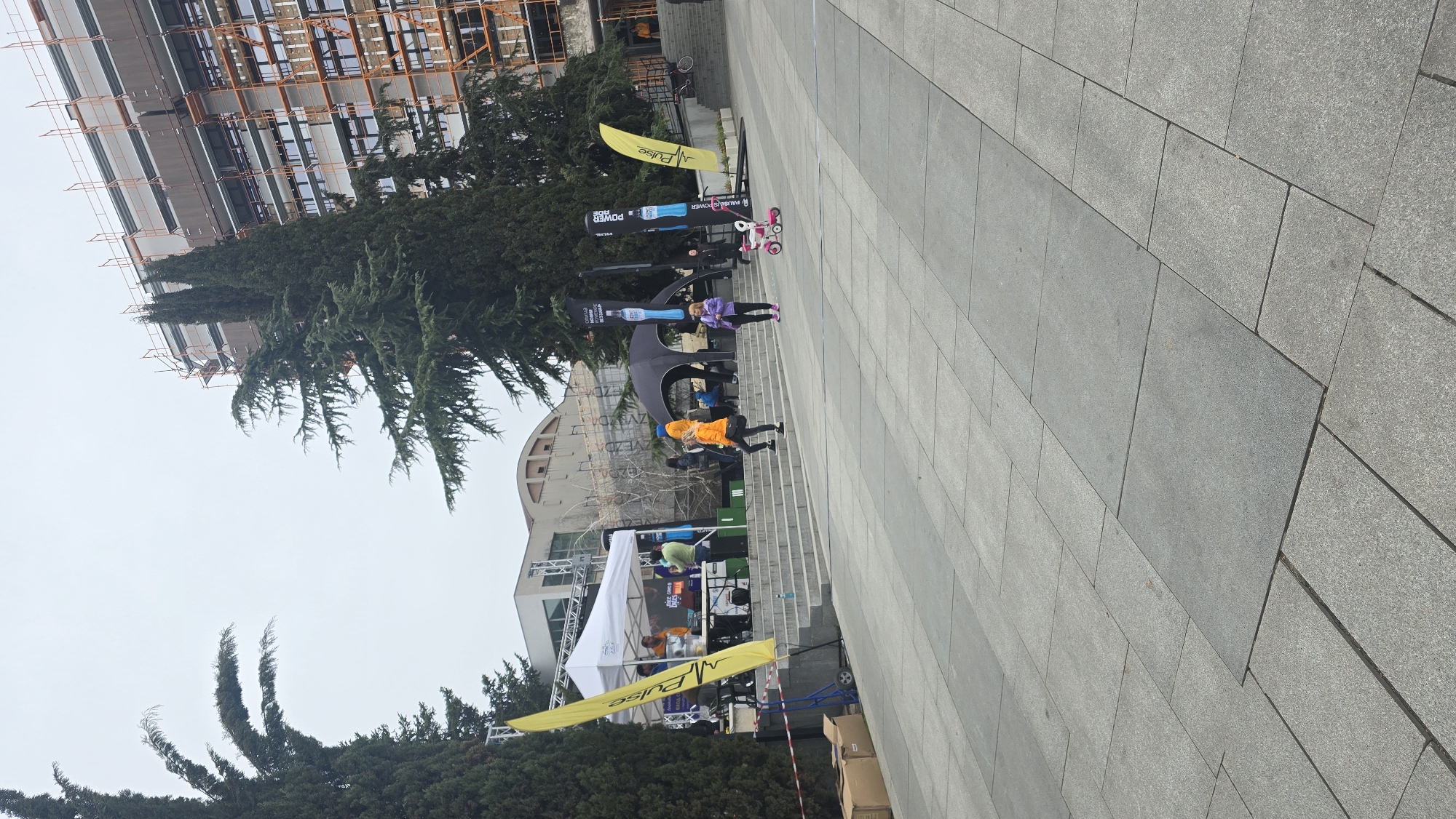 The image depicts an outdoor event taking place on a paved area in Stara Zagora, Bulgaria. A white tent is visible, suggesting a promotional or vendor space. People are observed walking and interacting around the tent and on the steps behind it. A large structure, possibly a building under construction or renovation, is partially visible in the background. Trees are present, and several yellow flags with the word "Pulse" on them are planted on the lawn. A black sculptural element is located in the background of the scene. Several promotional banners for "Powerade" are also visible. Several children are riding scooters.
