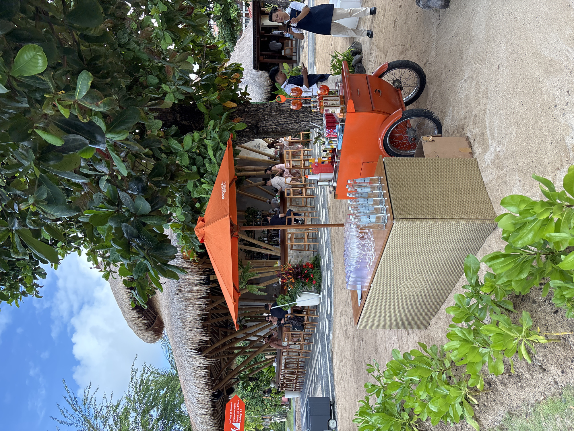 The image depicts an outdoor setting likely in Kuta Selatan, Indonesia, based on the tropical vegetation and architectural style. A thatched-roof structure houses a bar area with patrons seated at tables. An orange umbrella provides shade. A three-wheeled orange beverage cart stands nearby, stocked with bottles and glassware. Two individuals in waiter uniforms are visible tending to the cart. A rectangular table covered in a textured material also displays glassware. Sand covers the ground, and tropical foliage frames the scene. The sky is visible with some clouds.