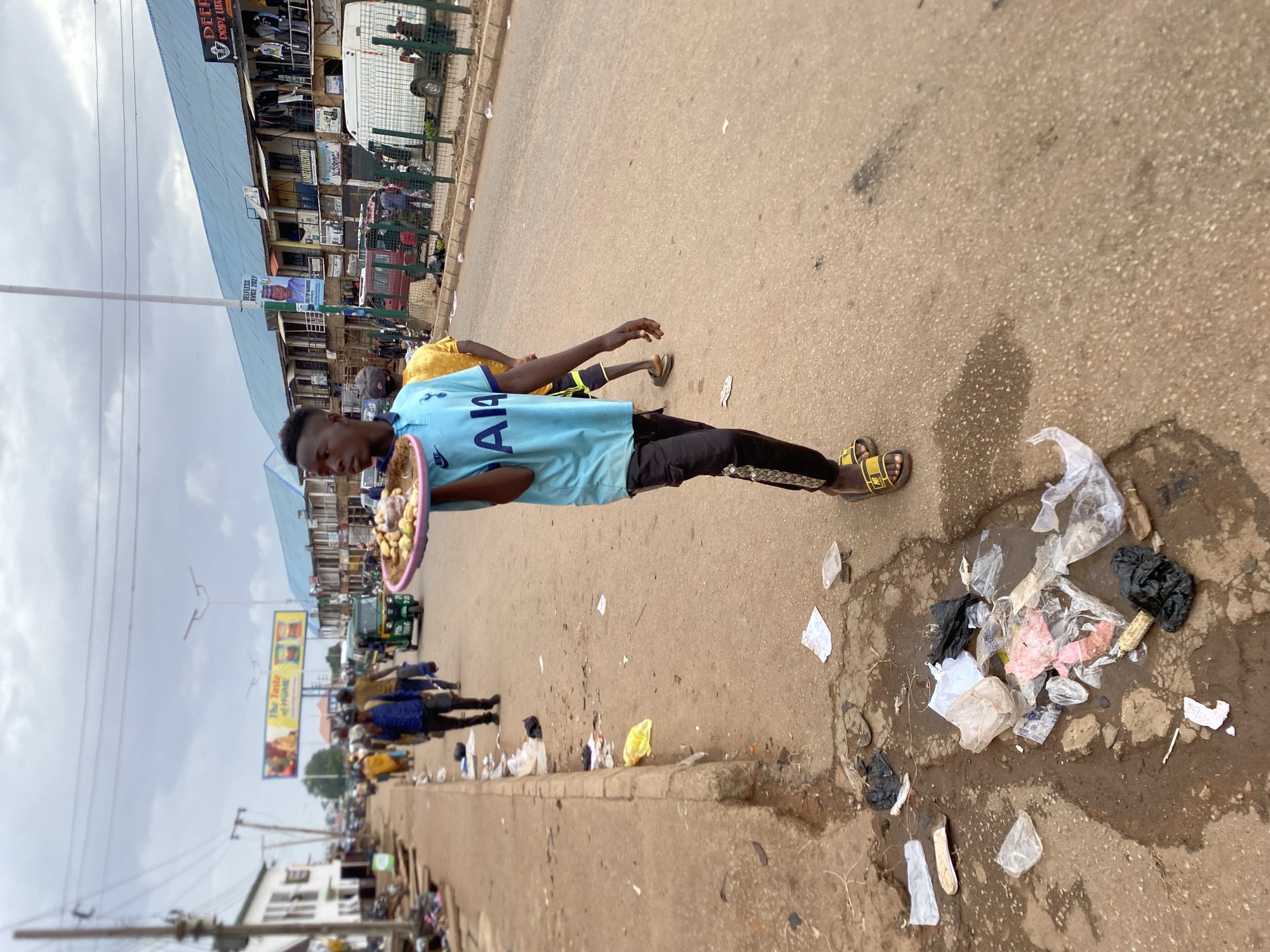 The image depicts a street scene in Bukuru, Nigeria. A young man wearing a light blue Tottenham Hotspur soccer jersey, black pants, and yellow sandals is carrying a pink tray laden with small, round fruits or snacks. He is walking along the side of a paved road.
Scattered along the roadside is a significant amount of litter, including plastic bags, paper, and other debris. In the background, there are buildings, signs, and other people walking. There is a three-wheeled vehicle that looks like a rickshaw. The sky is overcast.