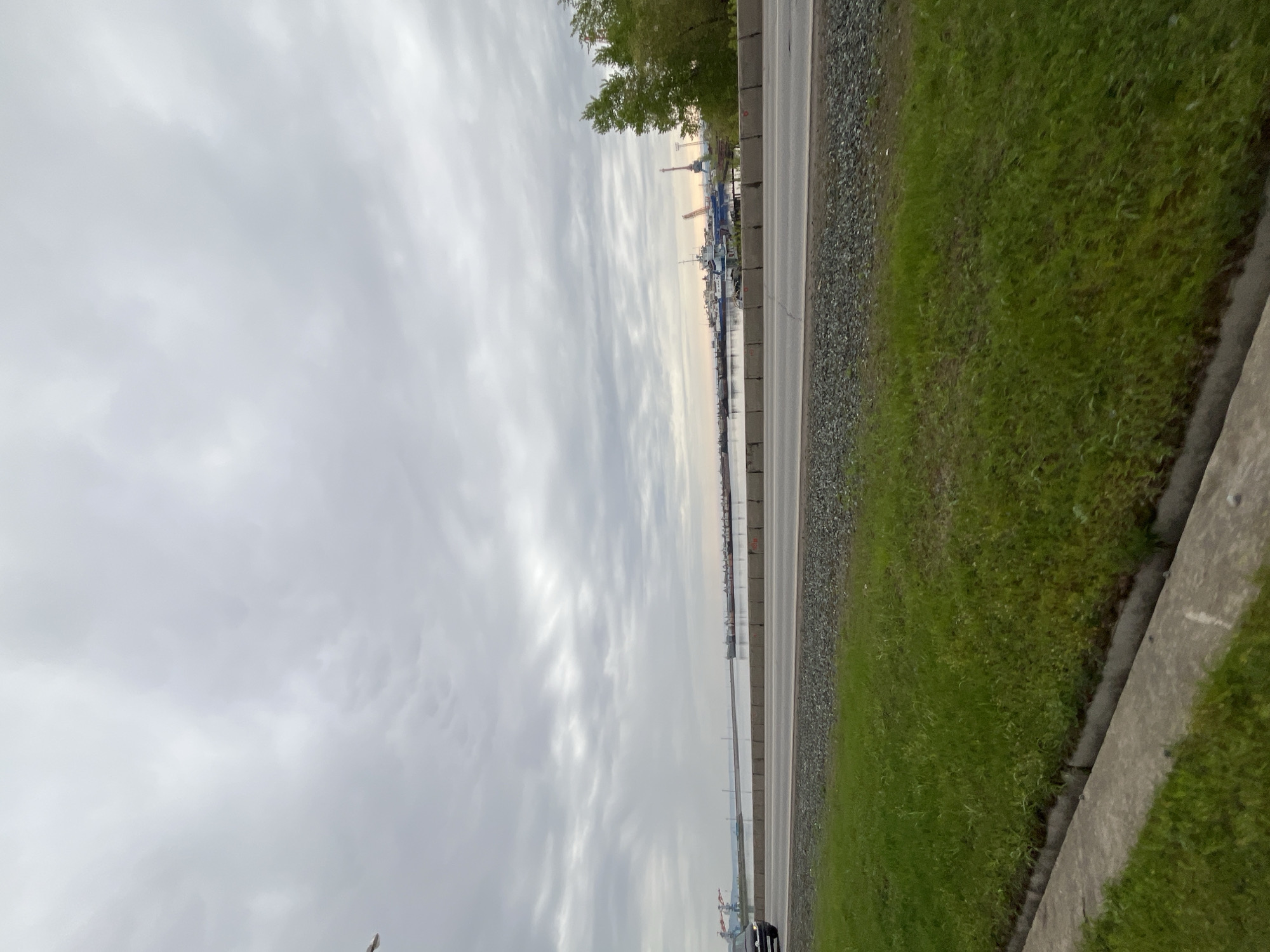 The image shows a view of the city of Chaikovsky, Russia, taken from a roadside perspective. A road runs horizontally through the center of the frame, flanked by green grass on one side and a body of water on the other. Beyond the water are industrial facilities, including cranes. The sky is overcast with layered clouds. A single car is visible on the road at the bottom edge of the frame.
