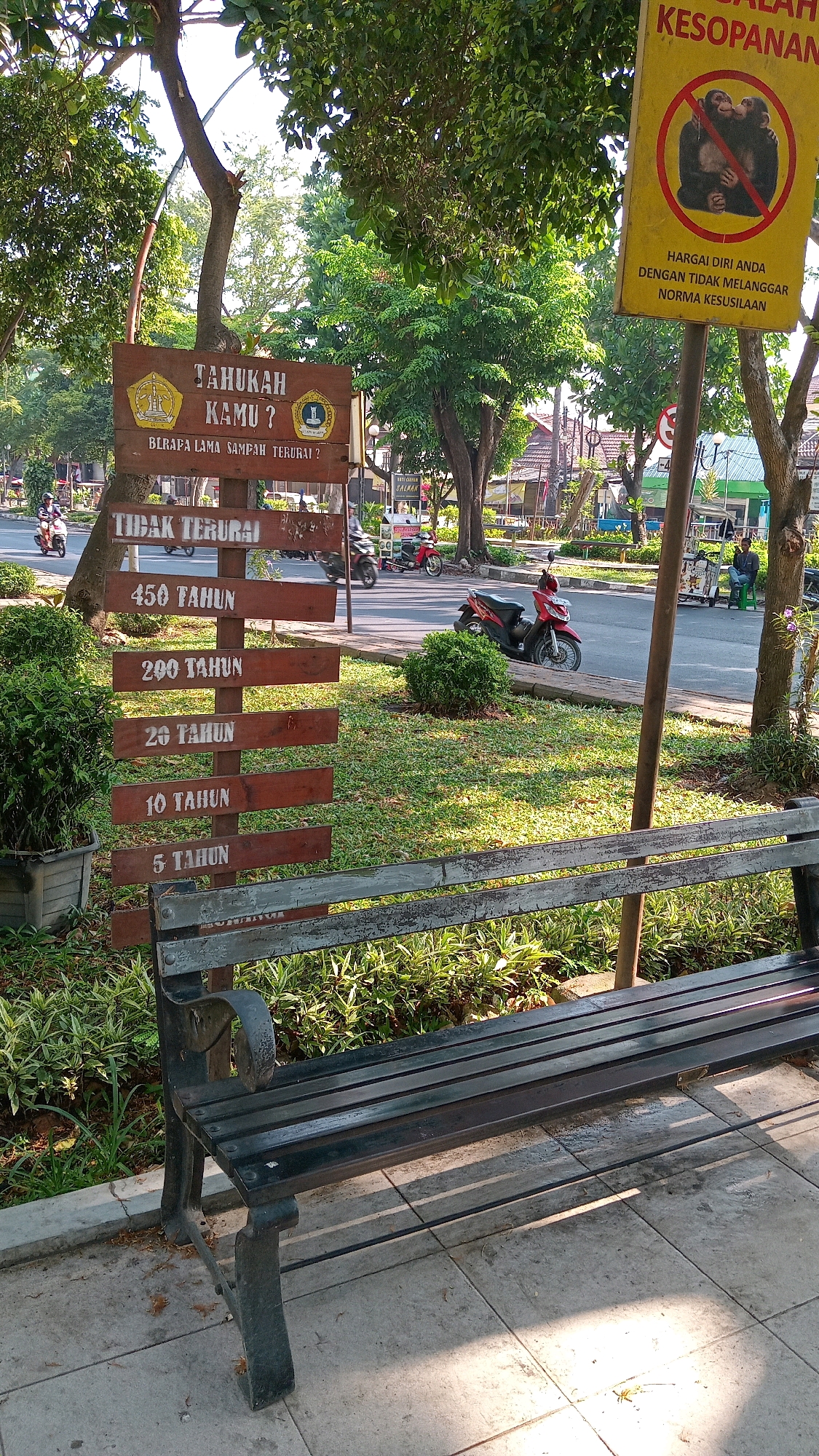 The image captures an outdoor scene in the city of Randuagung, Indonesia. In the foreground, a dark green park bench is positioned on a paved walkway, with a dense arrangement of green foliage behind it. To the left of the bench, a wooden signpost stands upright. This signpost features several horizontal wooden planks with text inscribed on them, likely indicating various decomposition times for different types of waste. The top planks read "TAHUKAH KAMU?" and "BERAPA LAMA SAMPAH TERURAI?". Below this, there is a plank that says "TIDAK TERURAI", followed by planks listing "450 TAHUN", "200 TAHUN", "20 TAHUN", "10 TAHUN", and "5 TAHUN". To the right of the signpost, a yellow sign with red circular border is attached to a pole. This sign displays an image of two monkeys and text in Indonesian related to etiquette or norms.
Behind the signpost and bench, a grassy area with scattered bushes leads to a street. Several motorcycles are visible on the street, some parked and others in motion, suggesting moderate traffic. Trees with lush green foliage line the street and the grassy area, casting shadows on the ground. In the background, buildings and more trees are discernible, contributing to the urban park-like setting. The overall lighting indicates it is daytime, with sunlight filtering through the trees.