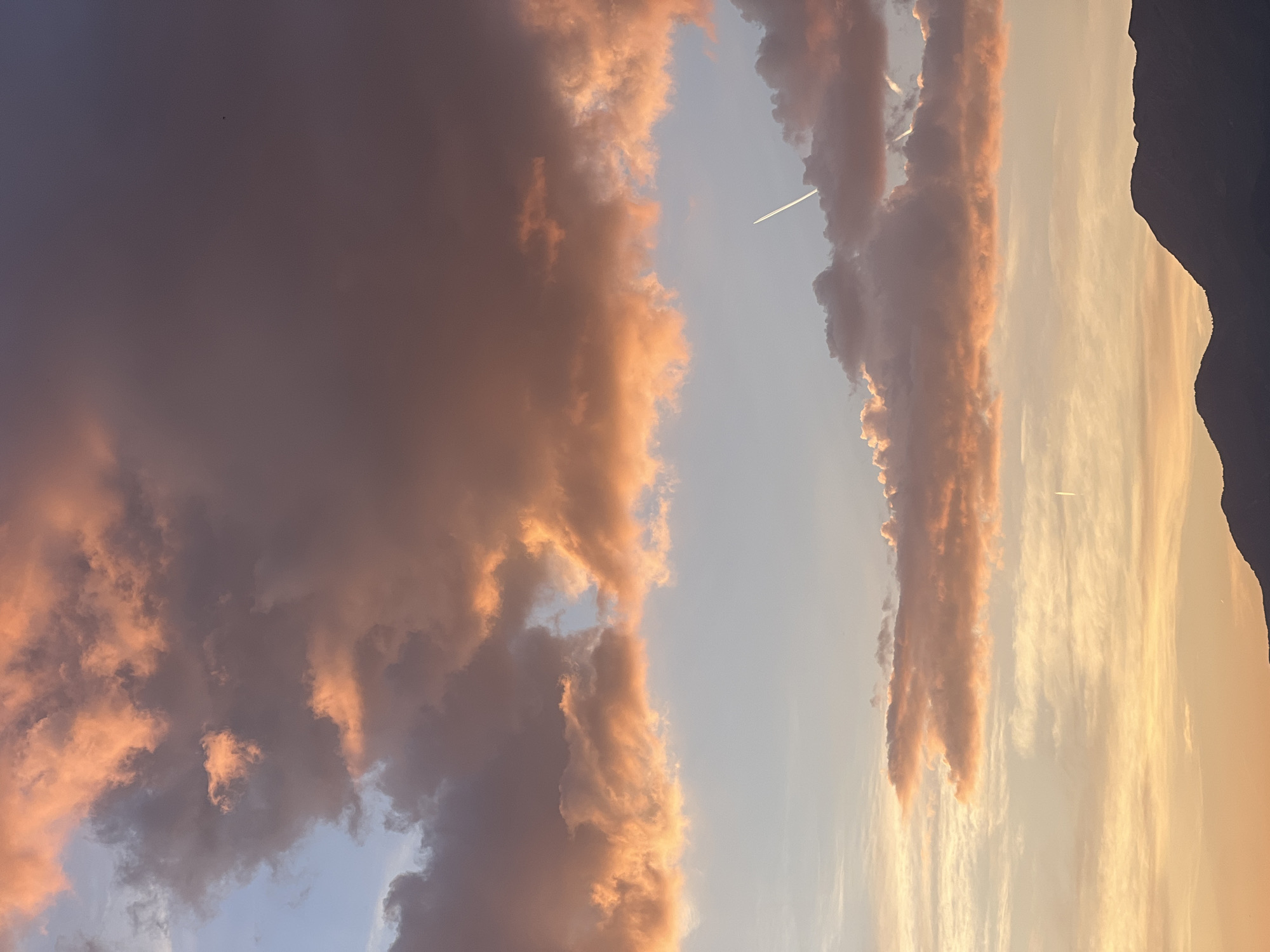 The image depicts a sunset sky over Sveti Stefan, Montenegro. There are orange and grey clouds in the sky, with a plane leaving a contrail. To the right, a dark silhouette of a mountain is visible. The overall impression is one of a serene evening.