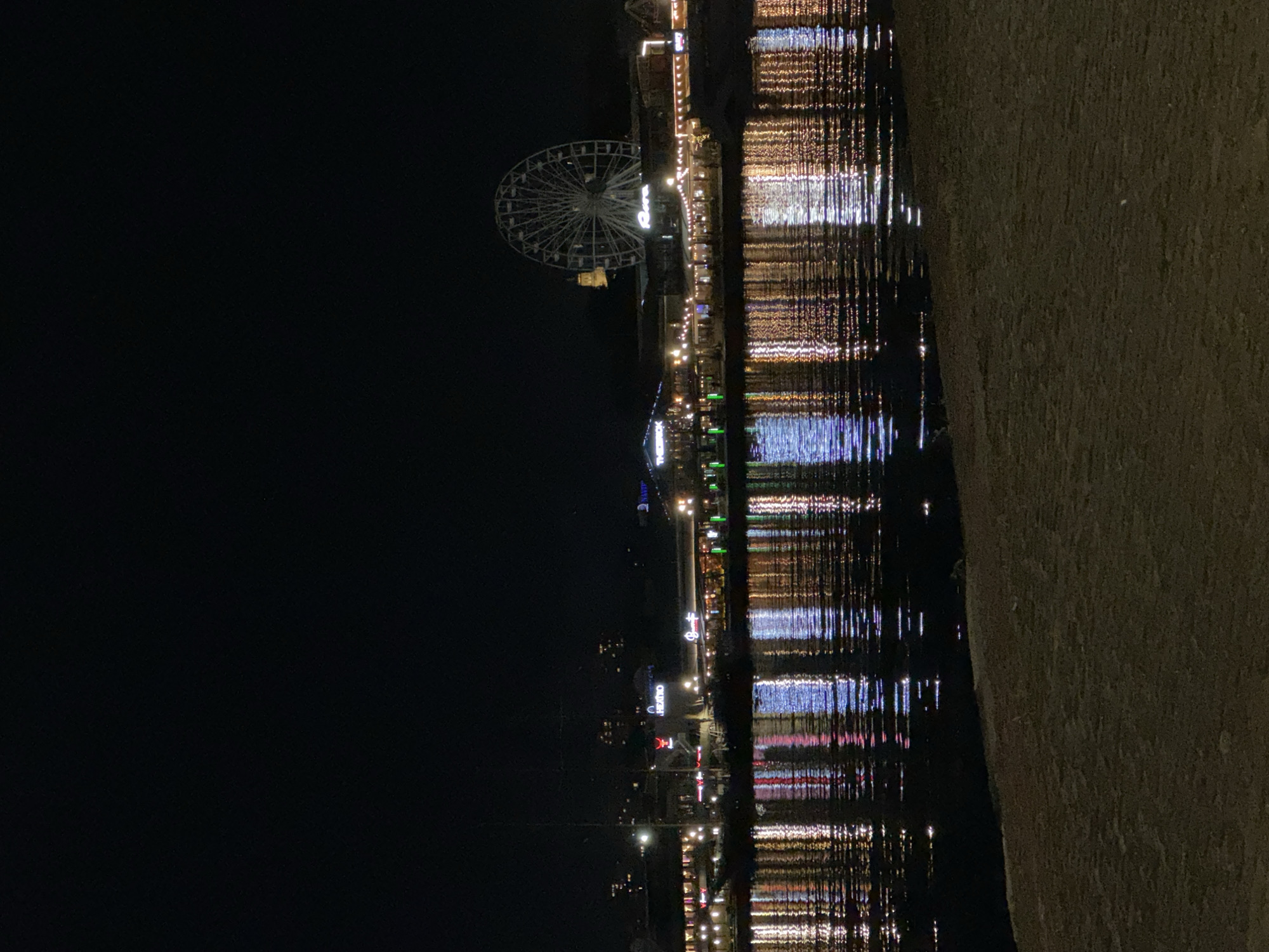 The image depicts the illuminated Varna waterfront at night, Bulgaria. A Ferris wheel is visible on the left. Lights from buildings and establishments reflect in the water, creating vertical streaks of white, blue, and orange. A stone structure borders the waterfront on the right.