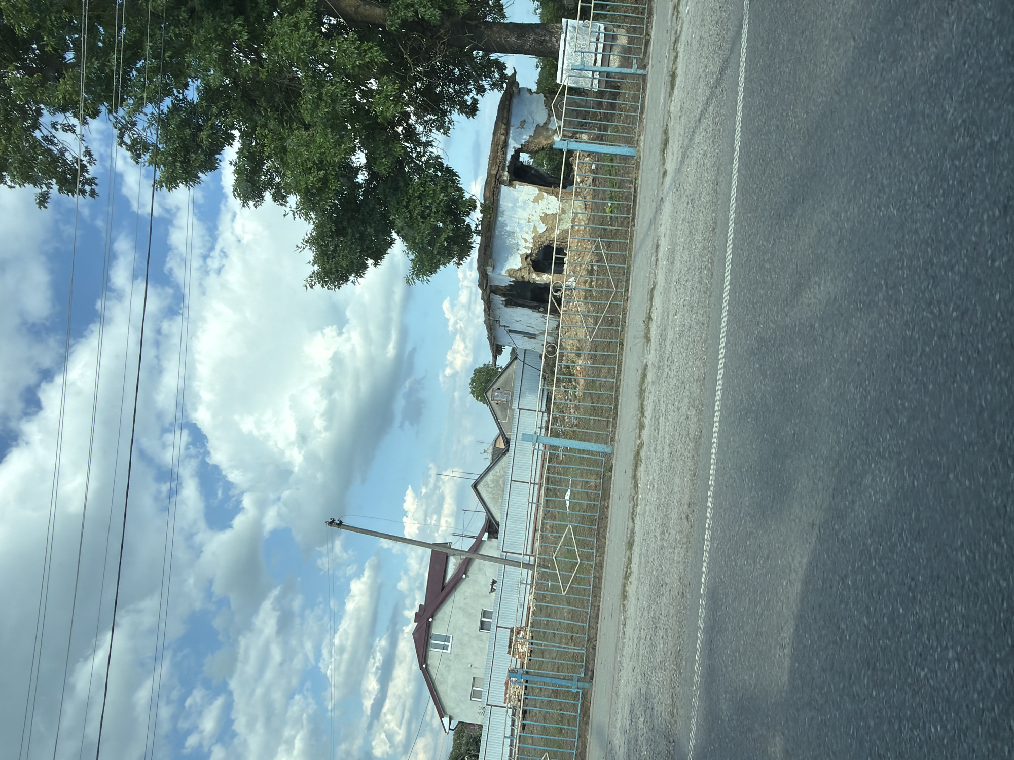 This media file depicts a roadside scene in Teofipilka, Ukraine, likely during the daytime under a cloudy sky. The image is oriented vertically.
The foreground is dominated by a paved road with a white dashed line marking its edge. To the left of the road, there is a gravel shoulder and then a blue metal fence. Beyond the fence, several structures are visible. A large, mature tree with lush green foliage stands prominently on the left, its branches extending over the fence and a damaged building. This building appears to be an old, partially collapsed structure with exposed walls and a gaping roof, suggesting it may be abandoned or has suffered damage.
Further down the fence line, there are houses with visible roofs and windows, one house being particularly prominent with a red-tiled roof and light-colored walls. Utility poles with overhead wires are also present, indicating the presence of infrastructure. The sky is a mix of blue and white clouds, suggesting a typical summer day. The overall impression is one of a rural or semi-rural setting with a contrast between newer residential buildings and older, decaying structures. There is no discernible activity or people in the image. No visible text is present.
